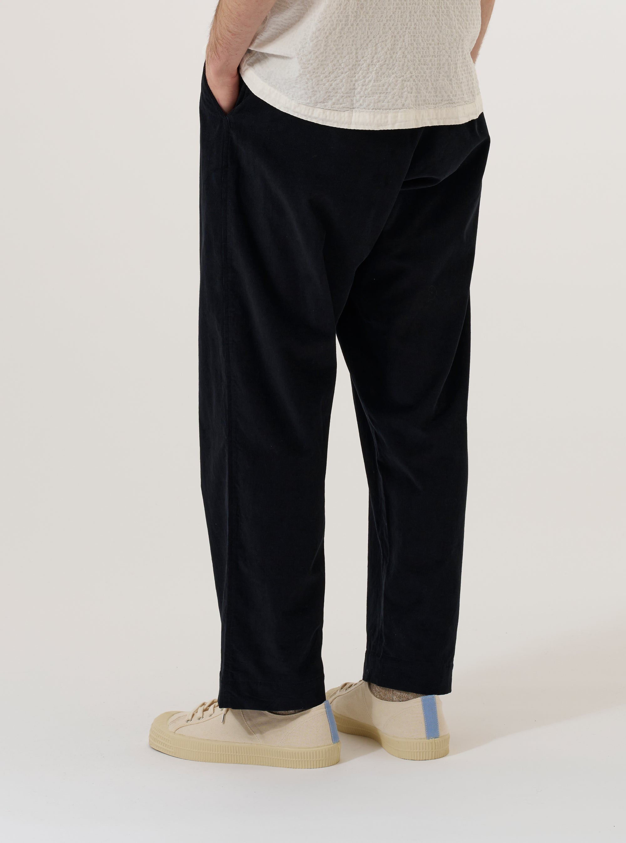 A model wears the Black Organic Fine Cord Oxford Pant, shown from the waist down against a plain, neutral background. The trousers feature a wide, loose leg with a single pleat front and are made from a black fine-wale corduroy fabric. The elasticated waistband with an external drawstring is clearly visible, and the model is standing with one hand in a front pocket, highlighting the garment's relaxed fit.