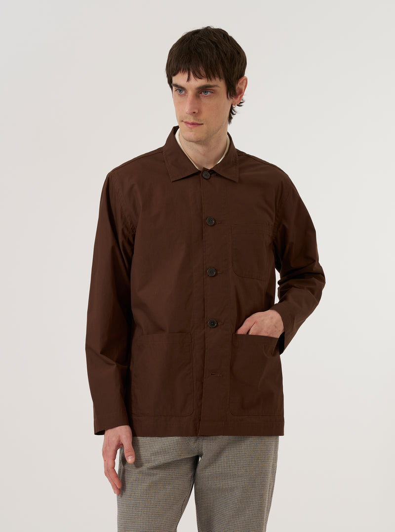A model wears the Brown Peached Poplin Bakers Overshirt over a light-colored shirt, standing against a plain white background. The overshirt features a classic collar, a five-button front closure, and multiple front patch pockets, including one at the chest and two at the waist. The cotton fabric has a smooth, matte peached finish in a rich brown color.