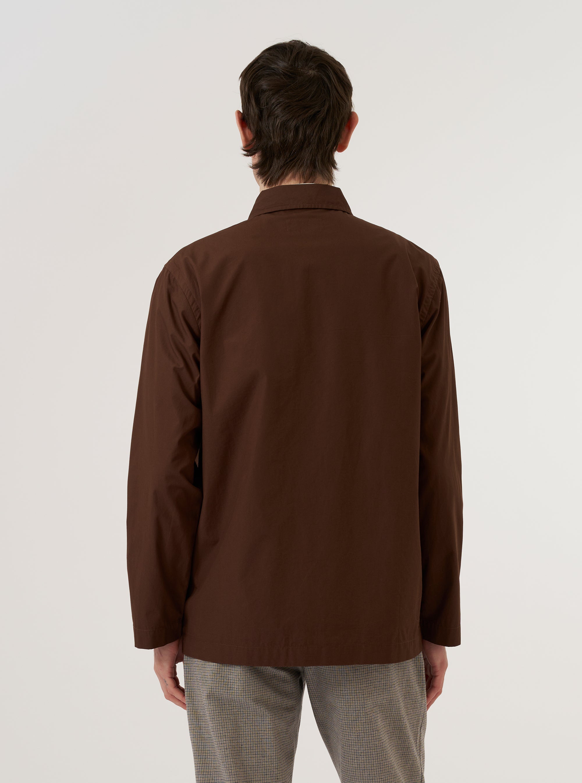 A model wears the Brown Peached Poplin Bakers Overshirt while standing against a plain white background. The image shows the garment from the back, highlighting its straight hem, relaxed boxy fit, and the smooth matte texture of the brown cotton poplin fabric. The model's hands are placed in the front pockets, partially visible from the side.