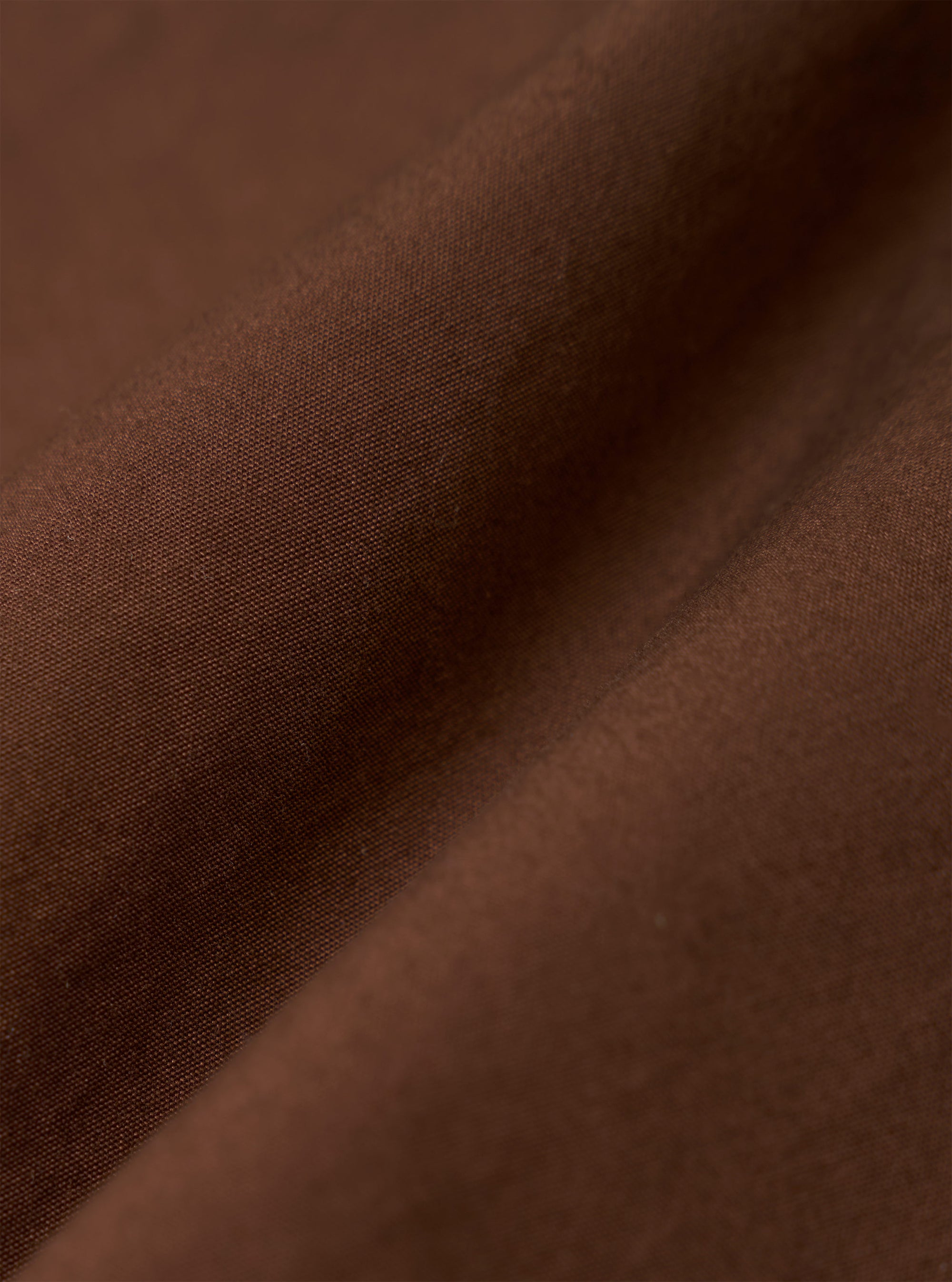 A close-up fabric swatch of the Brown Peached Poplin Bakers Overshirt is shown, highlighting the rich brown color and the smooth, soft texture of the organic cotton poplin fabric.
