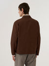 A model is wearing the Brown Peached Poplin Franck Jacket over a white t-shirt and light-colored trousers. The jacket is a brown, four-button blazer with three front patch pockets and a notched lapel. The model is standing against a plain white background, and the image captures the garment from the waist up.