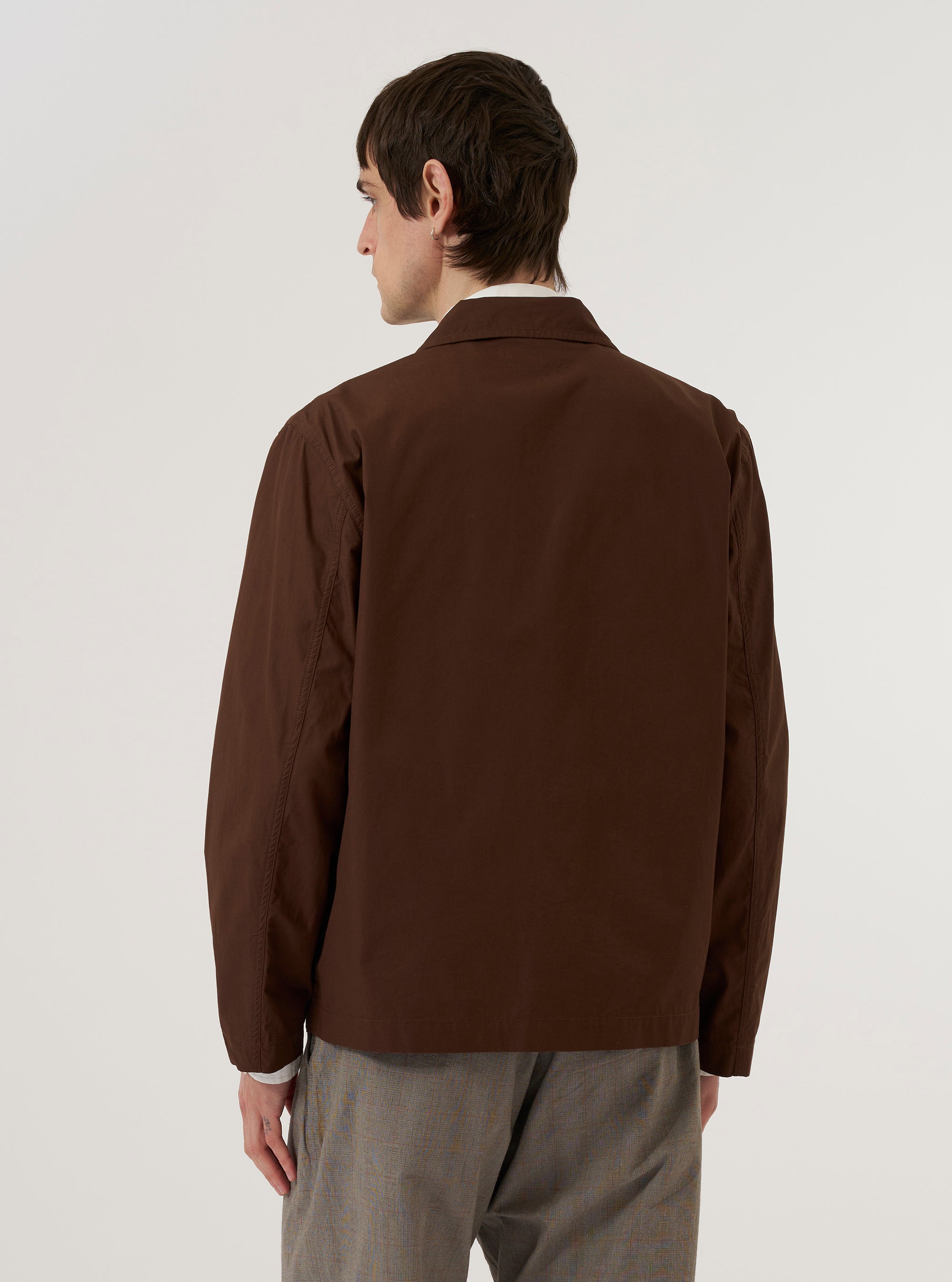 A model is wearing the Brown Peached Poplin Franck Jacket over a white t-shirt and light-colored trousers. The jacket is a brown, four-button blazer with three front patch pockets and a notched lapel. The model is standing against a plain white background, and the image captures the garment from the waist up.