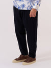 A model wears the Dark Navy Ospina Cotton Pleated Track Pant against a plain, neutral background. The image highlights the loose, tapered fit of the trousers, featuring a single pleat at the front and an elasticated waistband with a drawstring. The dark navy fabric has a visible seersucker texture.