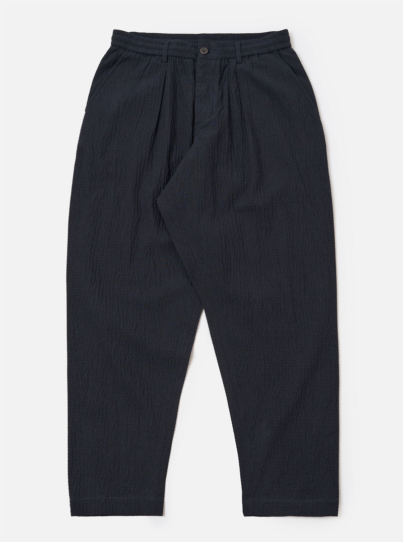 The Dark Navy Ospina Cotton Pleated Track Pant is shown as a flat lay against a plain white background. The trousers feature a single pleat front, an elasticated waistband with a matching drawstring, and a loose fit that tapers toward the hem. The dark navy fabric has a visible seersucker texture.