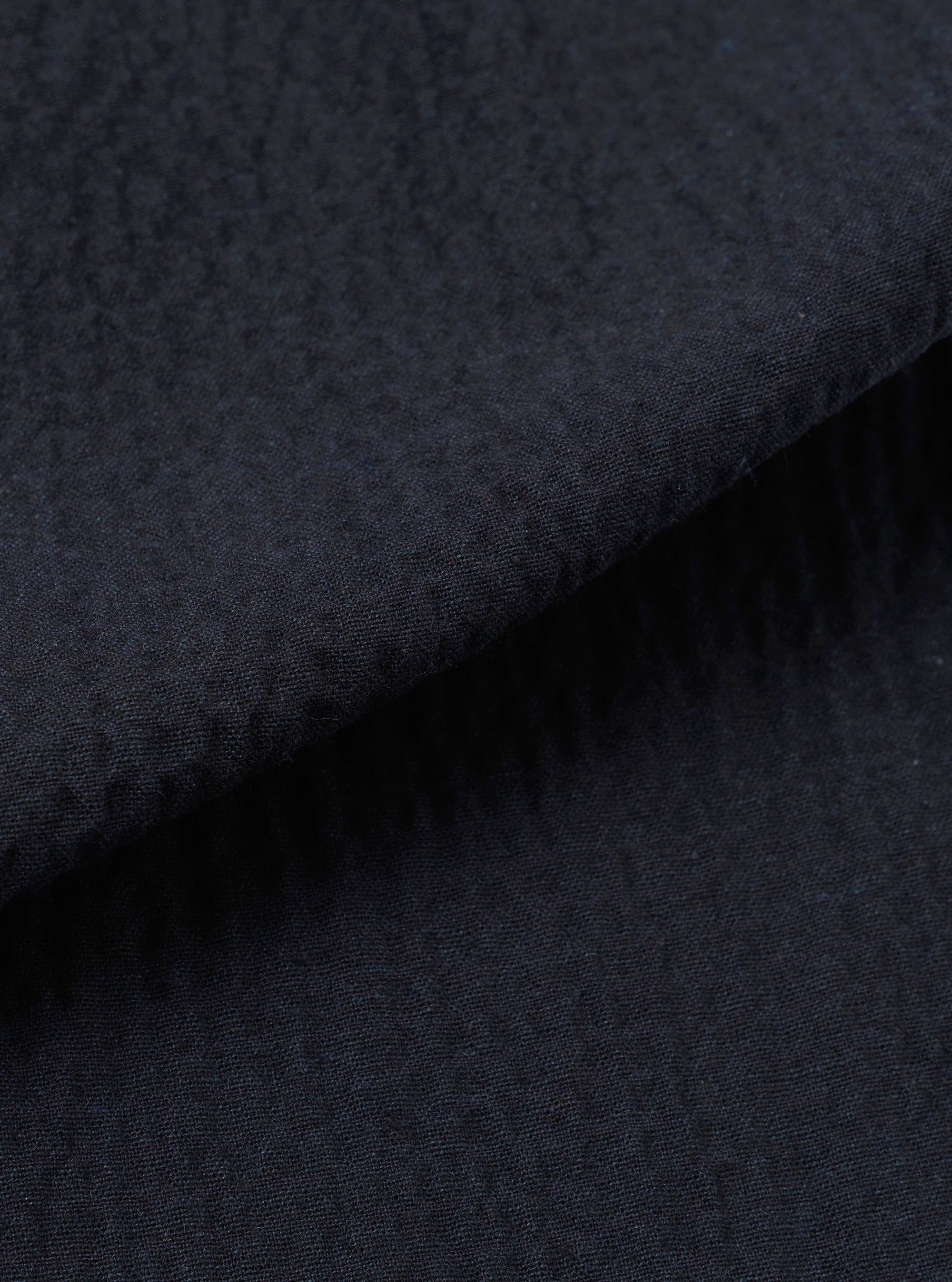 A close-up swatch of the Dark Navy Ospina Cotton Pleated Track Pant highlights the fabric's deep navy color and puckered seersucker texture. The material fills the frame, providing a detailed view of the recycled cotton blend's weave.