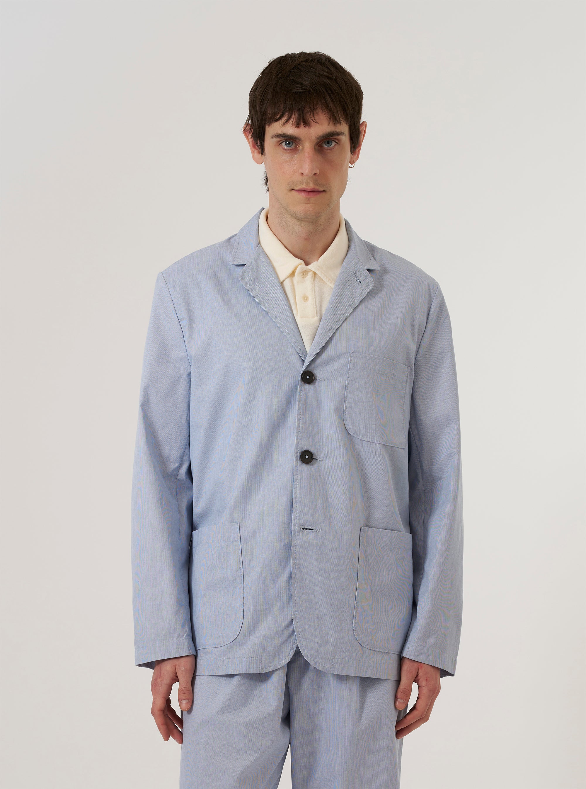 A model wears the Navy Kennedy Stripe Three Button Jacket over a light-colored shirt, standing against a plain white background. The jacket features a three-button front closure, three large patch pockets, and a classic lapel. The fabric has a vertical pinstripe pattern throughout.