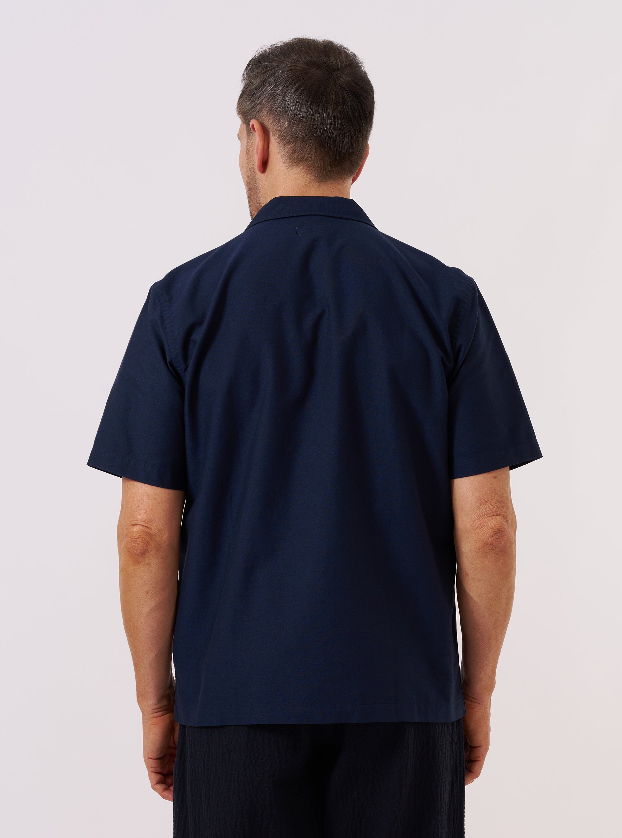 A model wears the Navy Classic Oxford Tropicana Shirt, shown from the back against a plain white background. The shirt features short, loose sleeves, a one-piece collar, and a straight-cut back with a slightly longer rear hem. The dark navy cotton fabric has a clean, matte finish. No mannequin is present in the image.