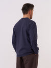 A model wears the Navy Recycled/Bio Jersey Henry L/S Tee, shown from the back against a plain white background. The long-sleeved garment features a relaxed fit and a straight hem, highlighting the soft texture and deep blue color of the recycled and organic cotton jersey fabric. No other accessories are prominent in this rear-view studio shot.