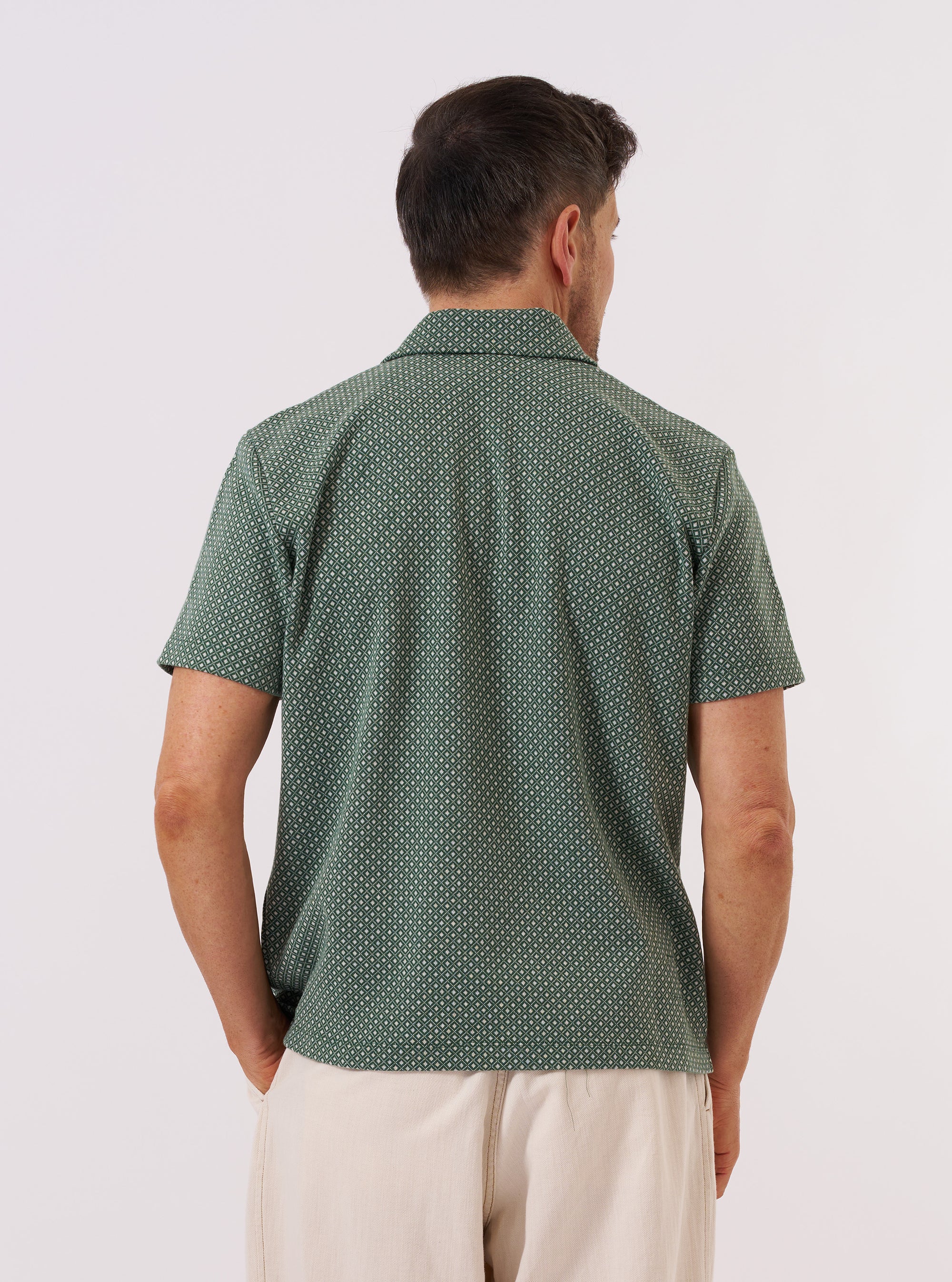 A model wears the Green Argyll Jersey Newlyn Polo against a plain white background. The polo features a short-sleeved design with a classic self-fabric collar, a two-button placket, and a large patch pocket on the left chest. The fabric is a green jersey with a tonal argyll diamond pattern throughout. The model is shown from the chest up, highlighting the collar and shoulder fit.