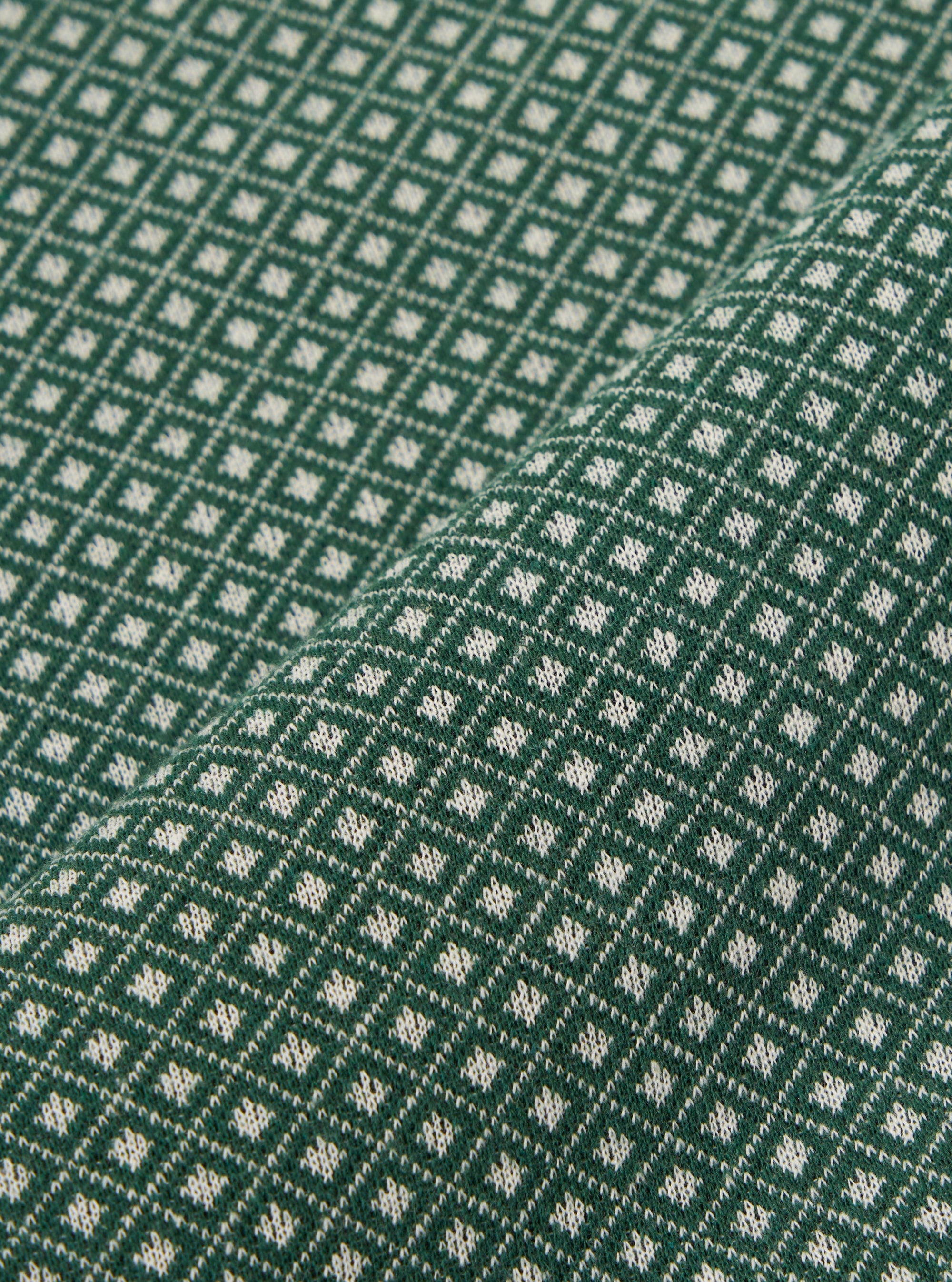 A close-up fabric swatch of the Green Argyll Jersey Newlyn Polo shows a green textile with a subtle, textured argyll pattern. The fabric is displayed flat, highlighting the intricate knit detail and varying shades of green.
