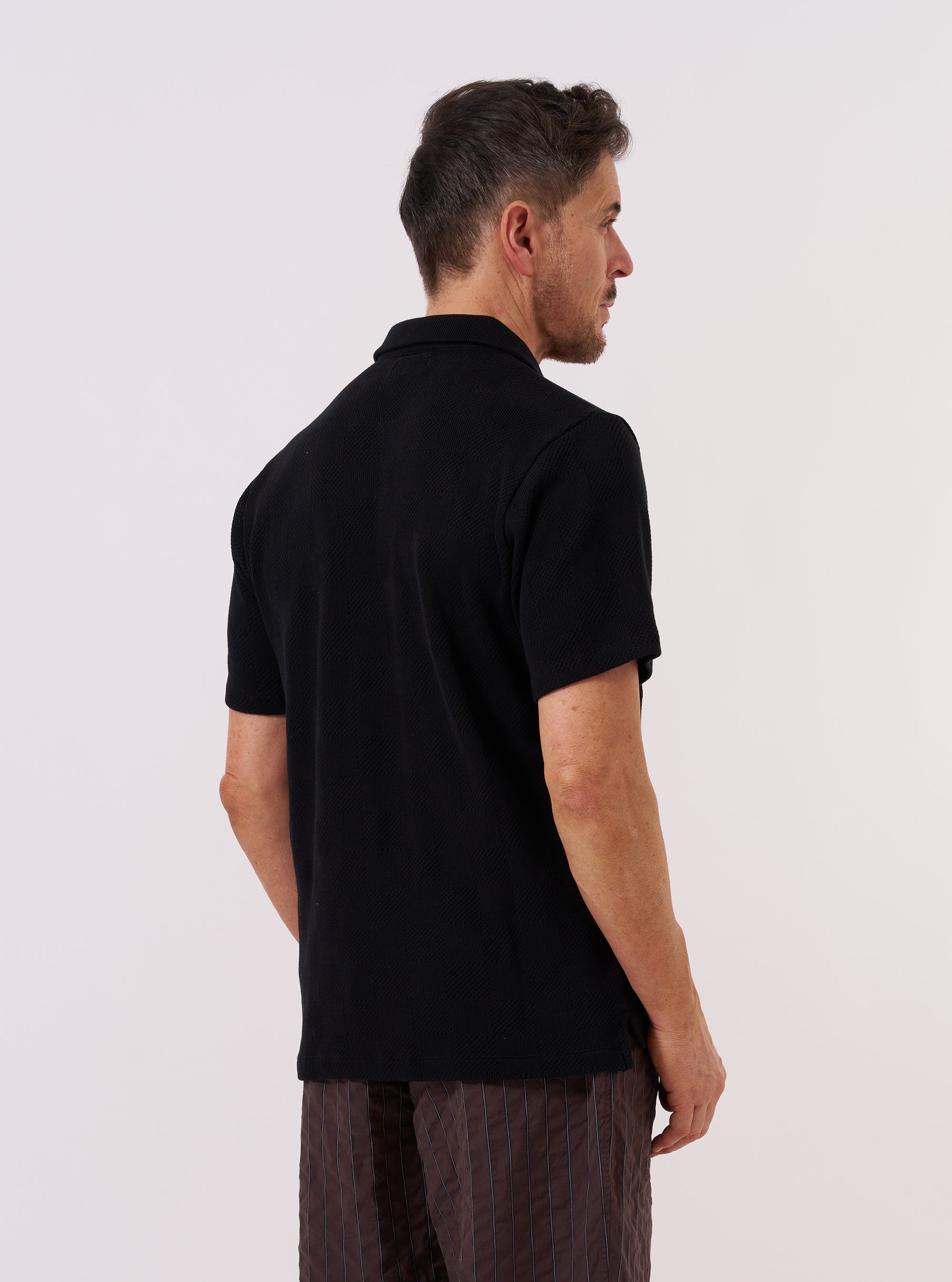 A model wears the Black Fantasy Checkerboard Vacation Polo, shown from a side angle against a plain, neutral studio background. The polo features an all-over black and grey checkerboard pattern, a relaxed fit, and a classic vacation collar. The textured organic cotton fabric and the short sleeves are prominent features in the image.