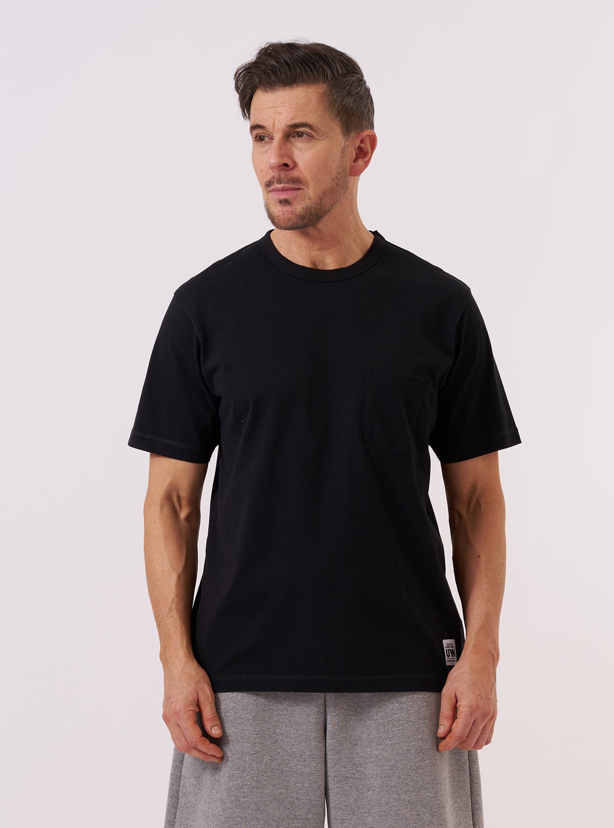 A model wears the Black Recycled/Bio Jersey Core Pocket Tee against a plain white background. The T-shirt features a classic crew neckline, short sleeves, and a single patch pocket on the left chest. The black jersey fabric has a smooth, matte finish. The model is shown from the chest up, highlighting the fit and the pocket detail.