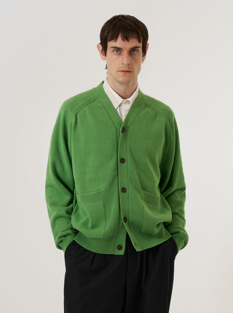 A model wears the Green Eco Cotton Knit Pocket David Cardigan over a light-colored shirt, standing against a plain white background. The cardigan features a V-neckline, a four-button front closure, and two large patch pockets at the waist. The green fabric has a visible knit texture.