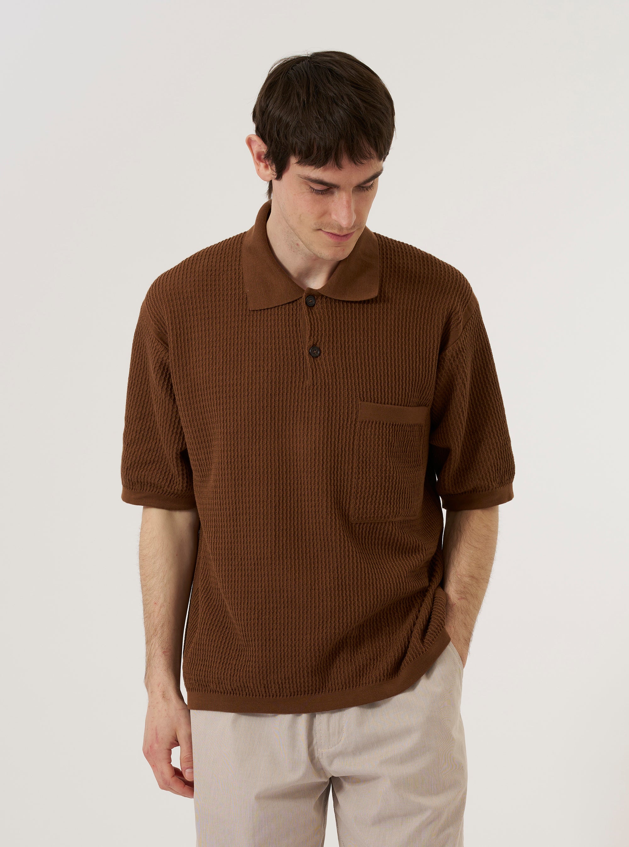 A model wears the Brown Fine Texture Tricot S/S Knit Polo against a plain white background. The polo features a short-sleeved design with a raised rib texture, a two-button placket, a neat collar, and a single square chest pocket. The model is shown from the chest up, highlighting the oversized fit and the intricate knit pattern of the brown cotton fabric.