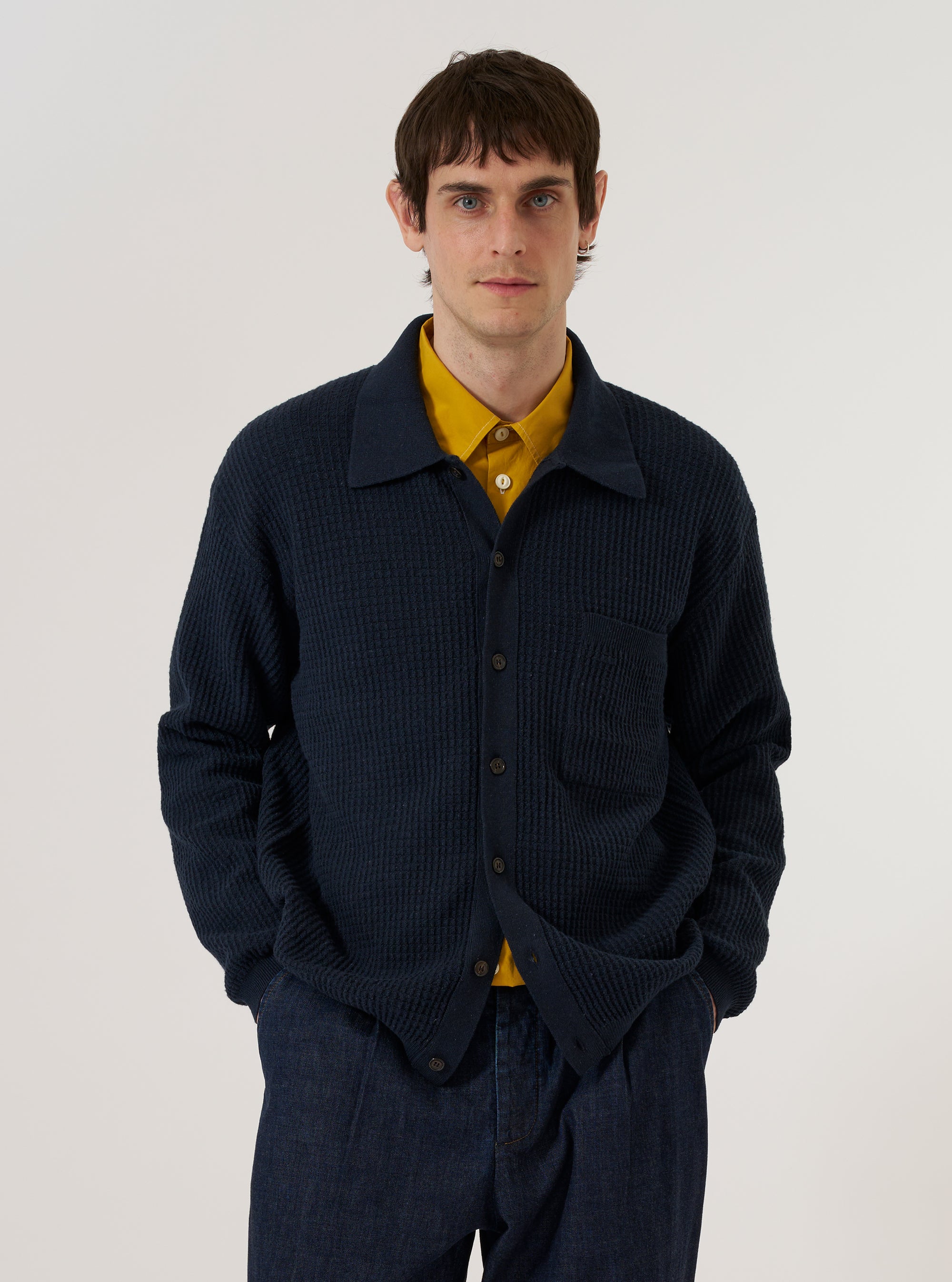 A model wears the Navy Eco Cotton Knit Jacket over a white t-shirt while standing against a plain white background. The jacket features a textured, chunky knit pattern, a classic button-up front with five buttons, and two lower patch pockets. The model is shown from the waist up in a front-facing studio shot.