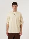 The Ecru Woolcot Bio S/S Pullover Knit Shirt II is worn by a model against a plain white background. The shirt features a classic collar, a three-button placket, and a single chest pocket. The short-sleeved garment is made from a textured ecru knit fabric and is worn tucked into dark trousers to highlight its relaxed fit.