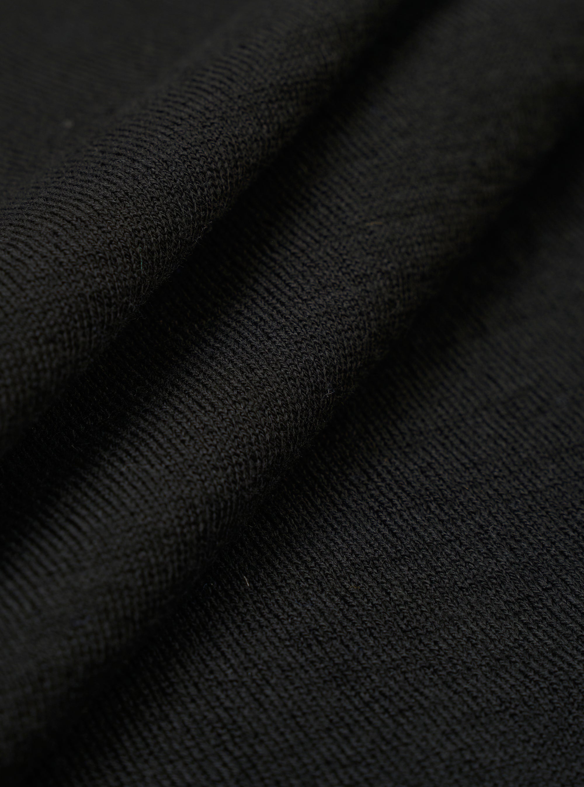 A close-up swatch of the Black Woolcot Bio S/S Pullover Knit Shirt II fabric highlights the smooth, fine-gauge texture of the black knit material. The image focuses on the blend of merino wool and organic cotton, with the fabric filling the entire frame.