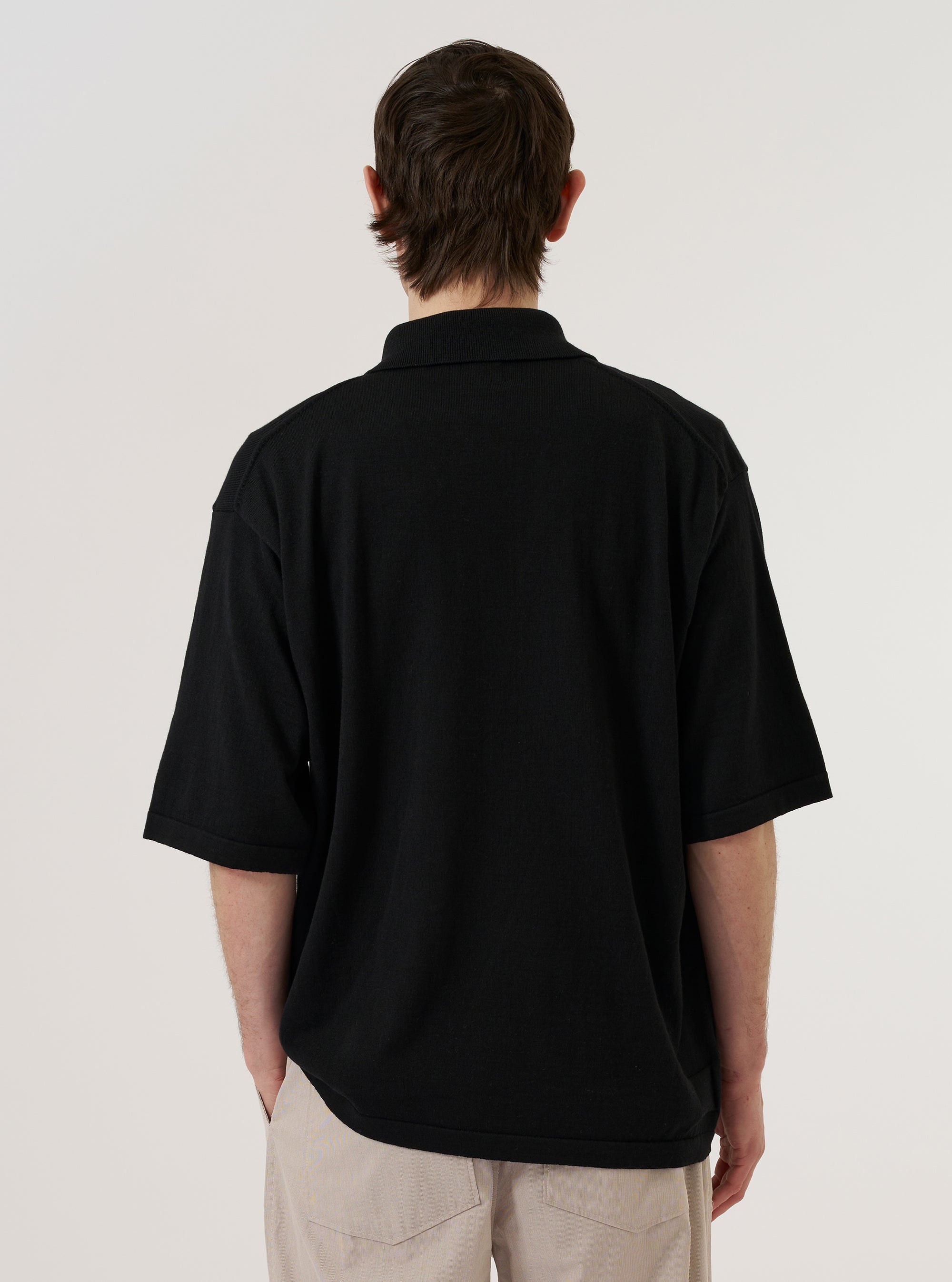 A model wears the Black Woolcot Bio S/S Pullover Knit Shirt II against a plain white background. The image, captured from the back at a slight angle, highlights the shirt's boxy fit, short sleeves, and smooth knit texture. The model is wearing the black pullover with light-colored trousers, showcasing the garment's relaxed silhouette and straight hem.