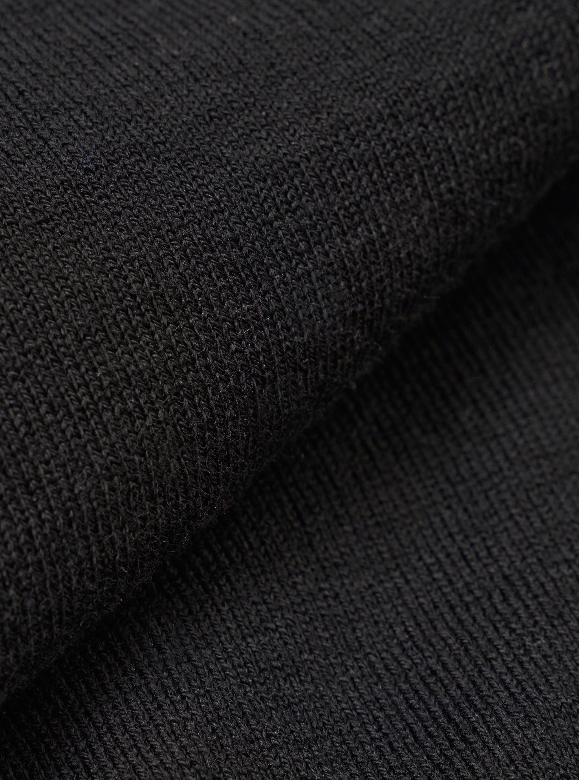 A close-up swatch image of the Black Woolcot Bio Loose Cardigan II fabric highlights the deep black color and the soft, fine-knit texture of the merino and organic cotton blend. The material fills the frame, providing a detailed view of the garment's knit construction.