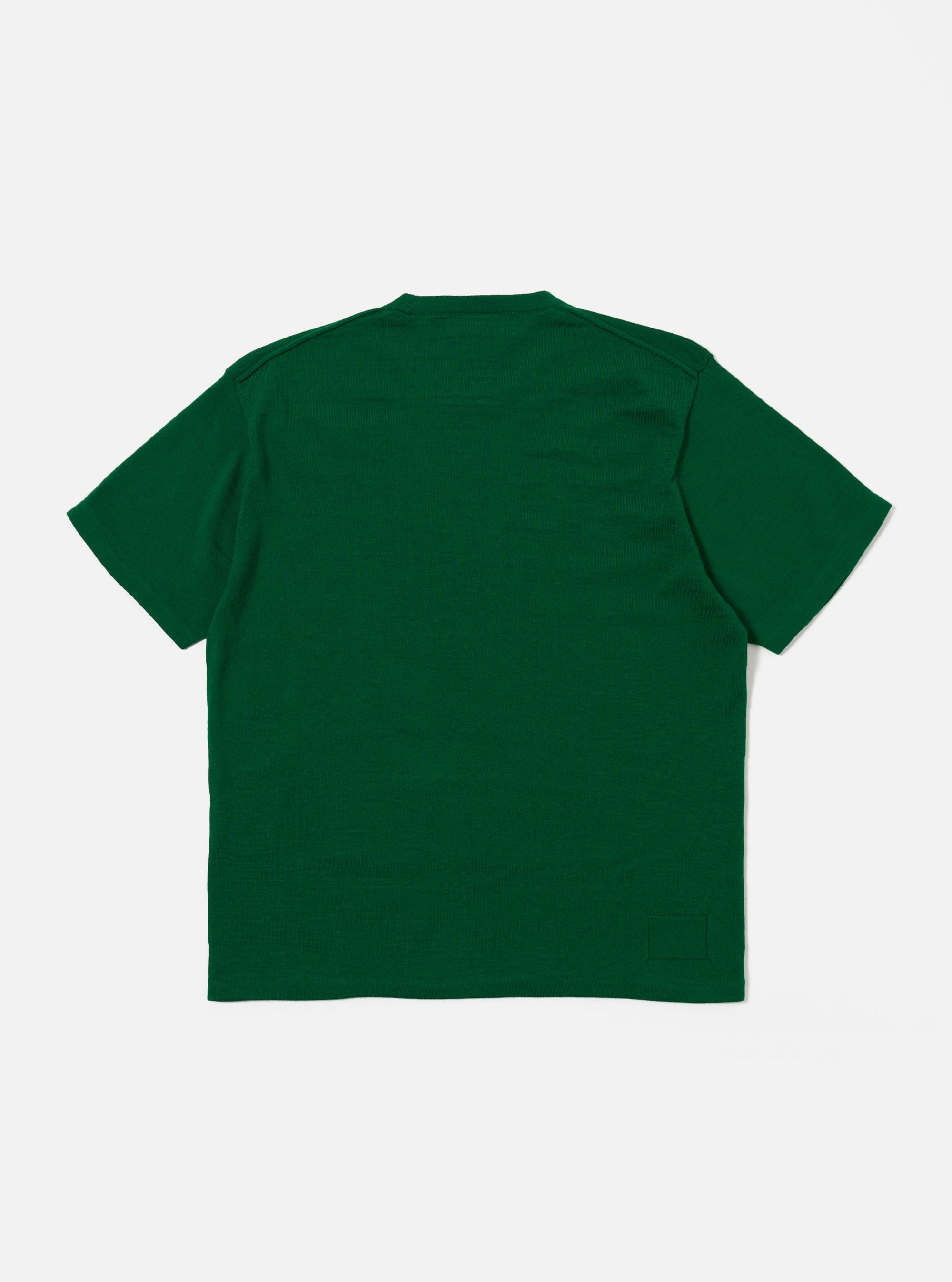 The Green Merino Fine Knit Knit Tee is shown from the back, worn by a model against a plain, light grey background. The image highlights the fine-gauge knit texture of the merino wool and the seamless fit across the shoulders and back. The model is also wearing dark trousers, but only the upper half of the body is visible. No other accessories are shown.