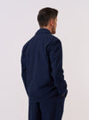 A model wears the Navy Derby Stripe Two Button Jacket over a light-colored top, shown from a side-angle view against a plain, neutral studio background. The jacket features a classic notched lapel, two-button front closure, and a visible vertical stripe pattern on the navy cotton fabric. The image highlights the tailored fit and the structured shoulder of the garment.