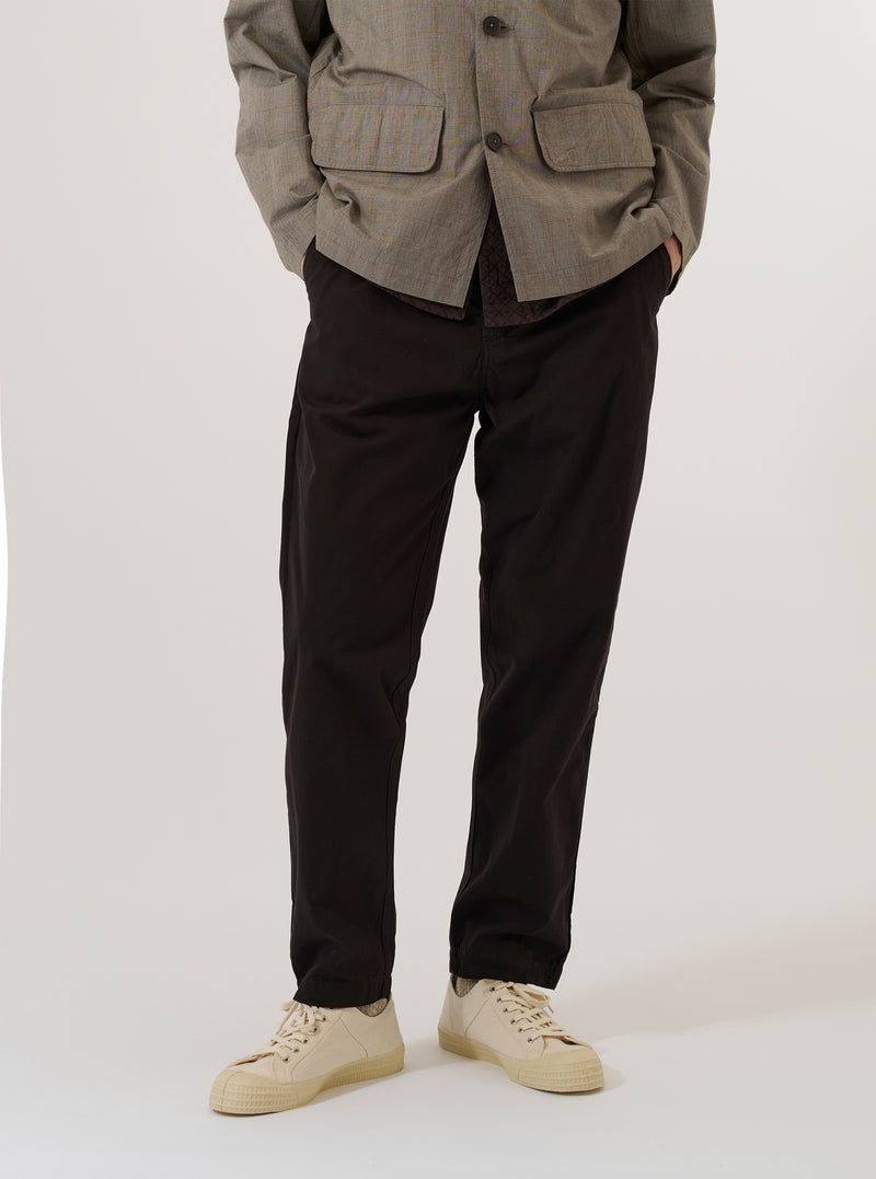 A model wears the Licorice Summer Canvas Military Chino while standing against a plain white background. The image shows the trousers from the waist down, highlighting the slim-straight fit, the dark brown cotton canvas fabric, and the classic chino styling with a button waist and side pockets. The model is also wearing white socks and brown loafers.