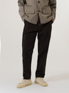 A model wears the Licorice Summer Canvas Military Chino while standing against a plain white background. The image shows the trousers from the waist down, highlighting the slim-straight fit, the dark brown cotton canvas fabric, and the classic chino styling with a button waist and side pockets. The model is also wearing white socks and brown loafers.