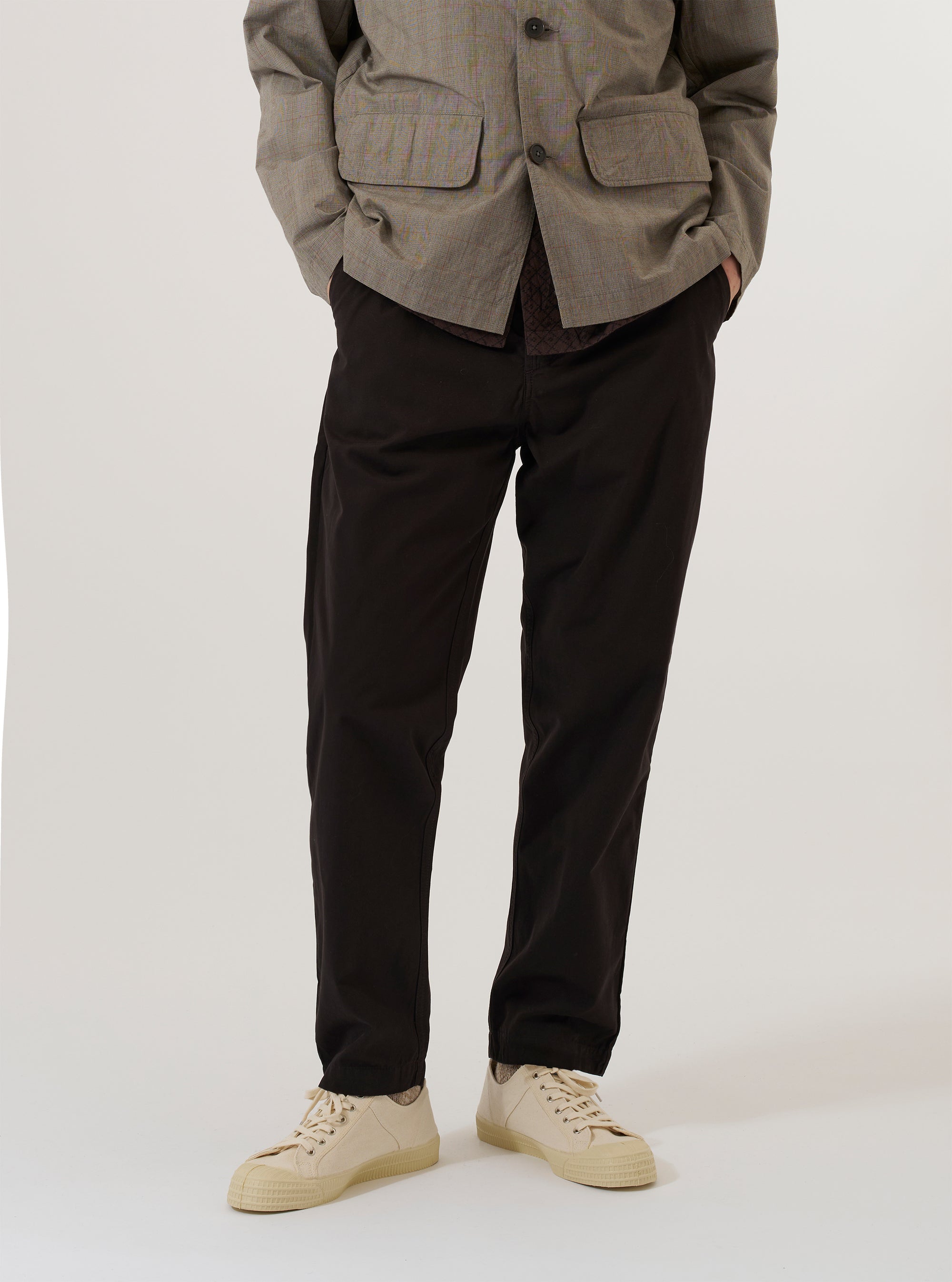 A model wears the Licorice Summer Canvas Military Chino while standing against a plain white background. The image shows the trousers from the waist down, highlighting the slim-straight fit, the dark brown cotton canvas fabric, and the classic chino styling with a button waist and side pockets. The model is also wearing white socks and brown loafers.