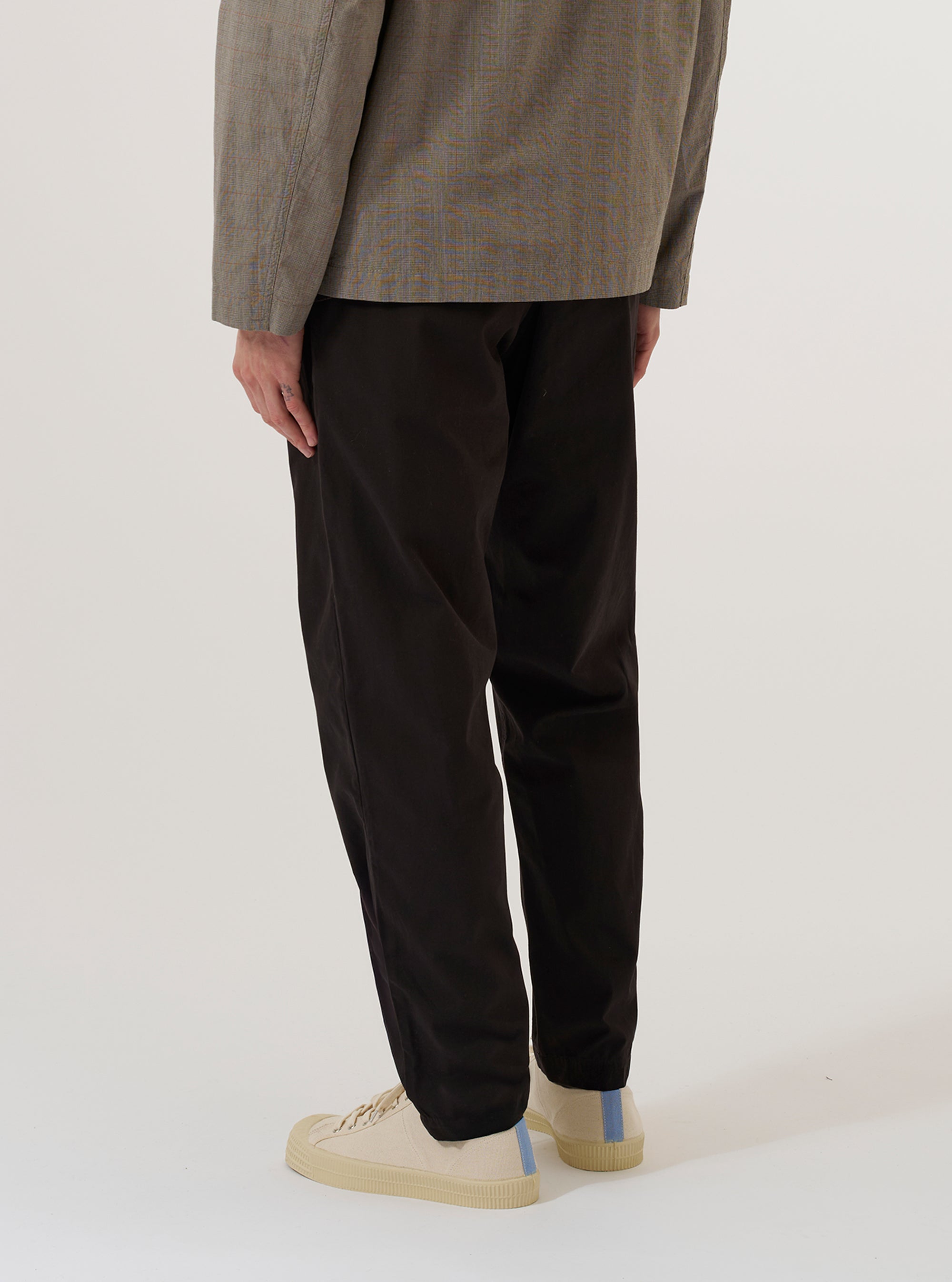 A model wears the Licorice Summer Canvas Military Chino, shown from the waist down against a plain white background. The trousers feature a straight-leg silhouette, side slant pockets, and a clean waistline. The garment is made from a dark licorice brown summer canvas fabric with a visible structured texture.