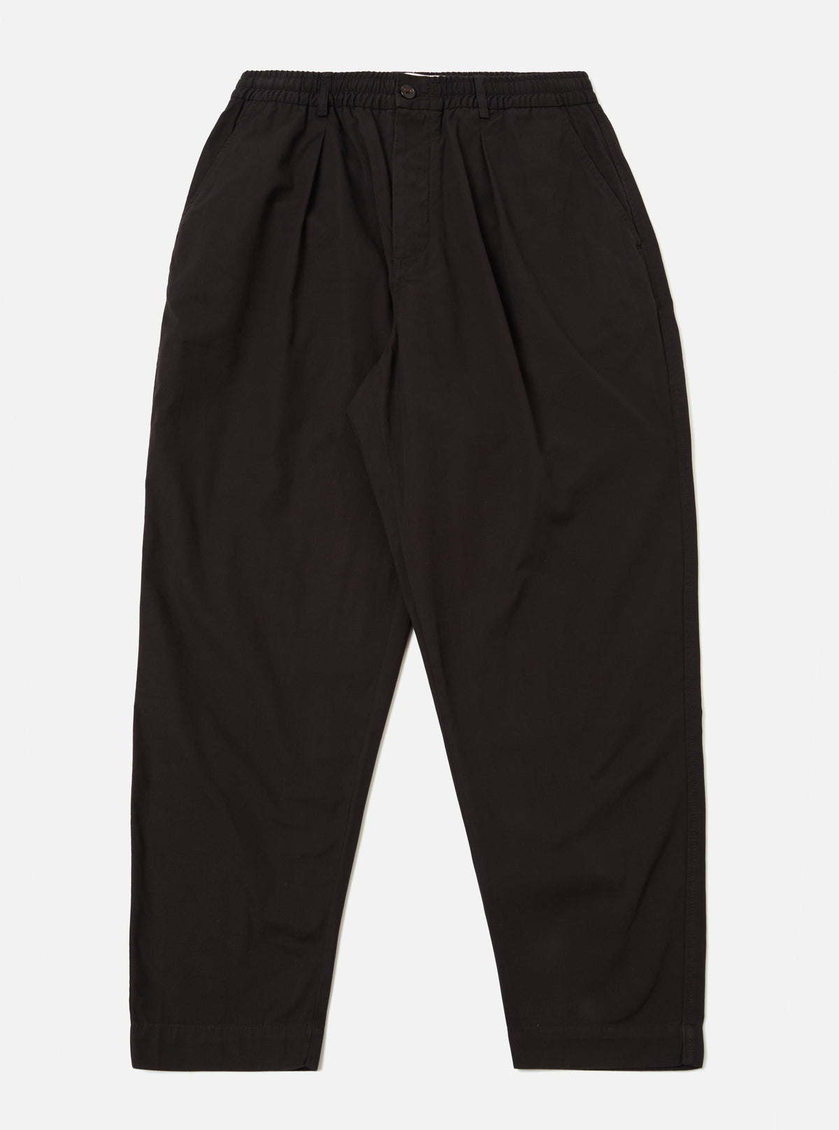 The Licorice Summer Canvas Pleated Track Pant is shown as a flat lay against a plain white background. The trousers feature an elasticated waistband with an internal drawstring, a single pleat on each leg, and a tapered silhouette. The fabric is a solid licorice brown cotton canvas with visible stitching at the hem and pockets.