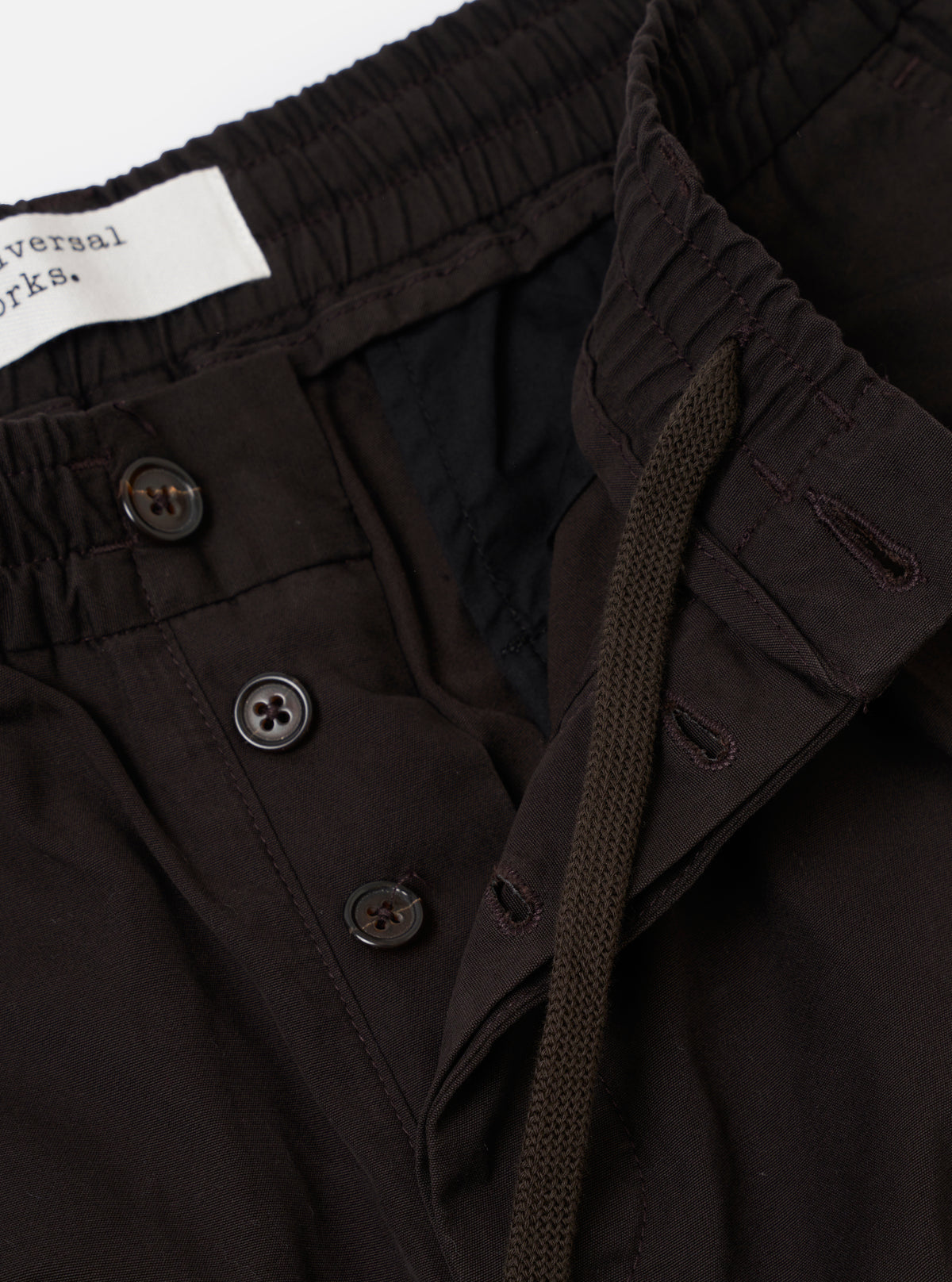 The Licorice Summer Canvas Pleated Track Pant is shown from the back as a flat lay against a plain white background. The image highlights the two rear pockets, one with a buttoned flap and one jetted with a button closure, along with the elasticated waistband and the textured, dark licorice-colored cotton canvas fabric.