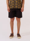 A model wears the Licorice Summer Canvas Clean Short, shown from the waist down against a plain white background. The shorts feature a relaxed fit, a mid-thigh length, and are made from a dark brown cotton canvas fabric. The model is wearing the shorts with white socks and brown shoes.