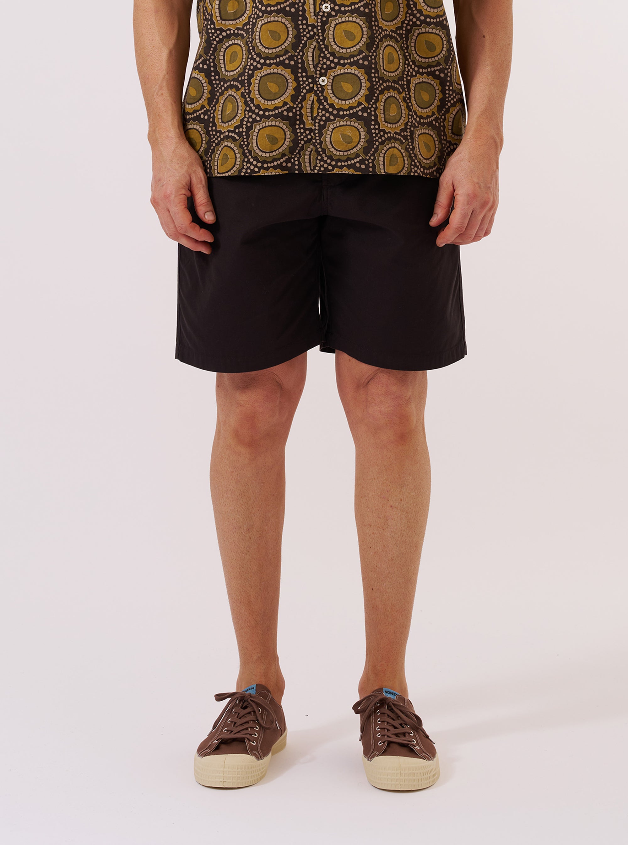 A model wears the Licorice Summer Canvas Clean Short, shown from the waist down against a plain white background. The shorts feature a relaxed fit, a mid-thigh length, and are made from a dark brown cotton canvas fabric. The model is wearing the shorts with white socks and brown shoes.