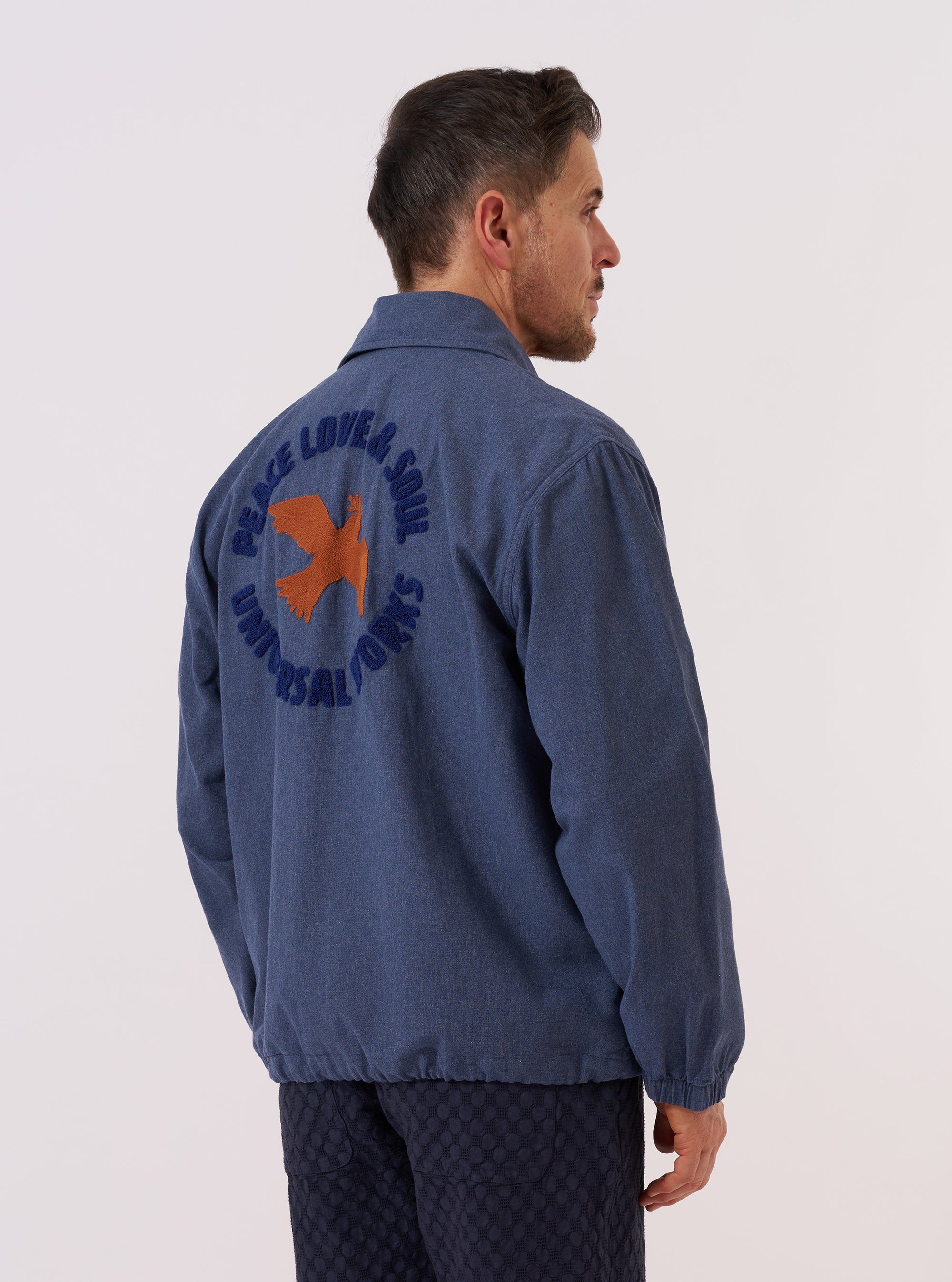 A model wears the Blue Sintra Recycled Cotton Head Coach Jacket over a light-colored top, shown from a side-angle view against a plain white background. The jacket features a classic pointed collar, a central button-down closure, and a straight hem. The garment is constructed from a blue recycled cotton and polyester blend fabric with a smooth, durable finish.