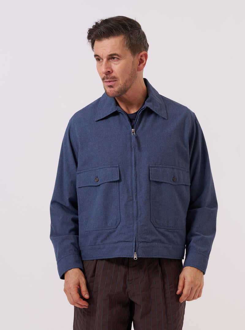 A model wears the Blue Sintra Recycled Cotton E130 Jacket over a light-colored shirt, standing against a plain, neutral background. The jacket features a relaxed, boxy fit, a two-way zip front, and two large front button-flap pockets. The light blue fabric has a visible textured finish with reinforced elbow patches and adjustable button cuffs.