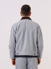 A model wears the Light Indigo Jean Cloth Reworked Rose Bowl Jacket, shown from a rear three-quarter angle against a plain white background. The jacket features a relaxed fit with a straight hem, a structured collar, and long sleeves. The light indigo organic cotton fabric has a visible textured grain, and the back is constructed with a clean, one-piece panel. No mannequin is present in the image.