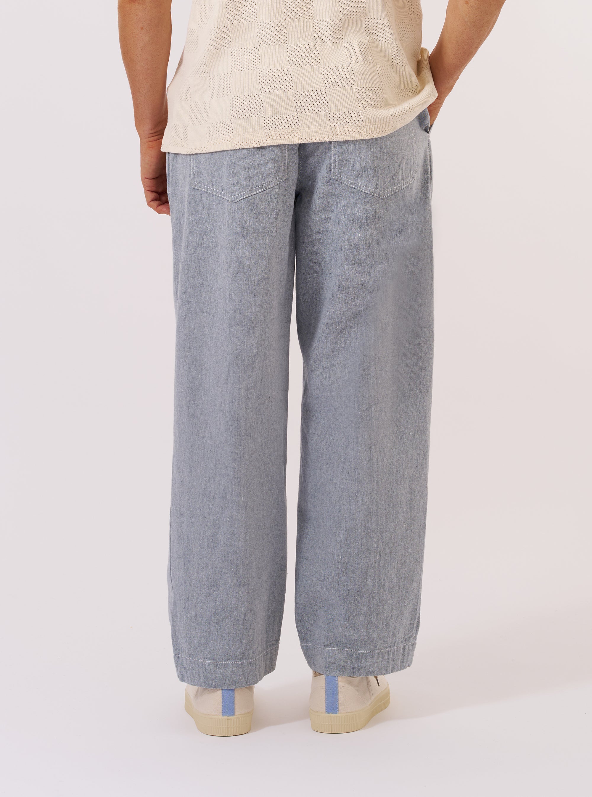 A model wears the Light Indigo Jean Cloth Reworked Duke Pant, shown from the back against a plain white background. The trousers feature a wide, loose fit, two large rear patch pockets, and a visible hammer loop on the left leg. The light indigo organic cotton fabric has a textured, washed appearance. The model is wearing the trousers with white socks and dark shoes.