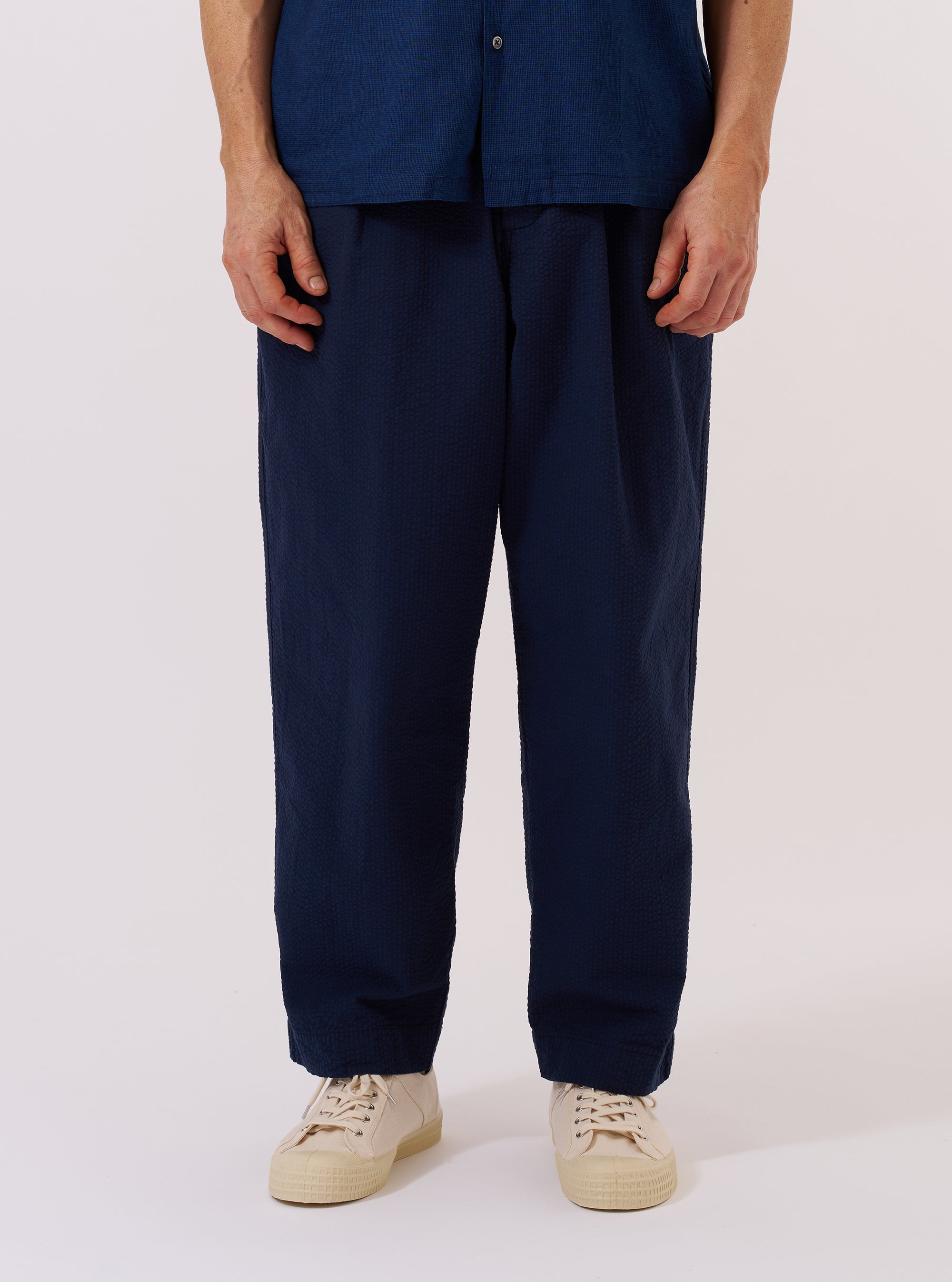A model wears the Navy Derby Stripe Oxford Pant against a plain white background. The trousers feature a loose, wide-leg fit with a pleated front and a drawstring waistband. The navy blue cotton fabric has a prominent vertical white stripe pattern throughout. The model is shown from the waist down, paired with a light-colored shirt and dark shoes.