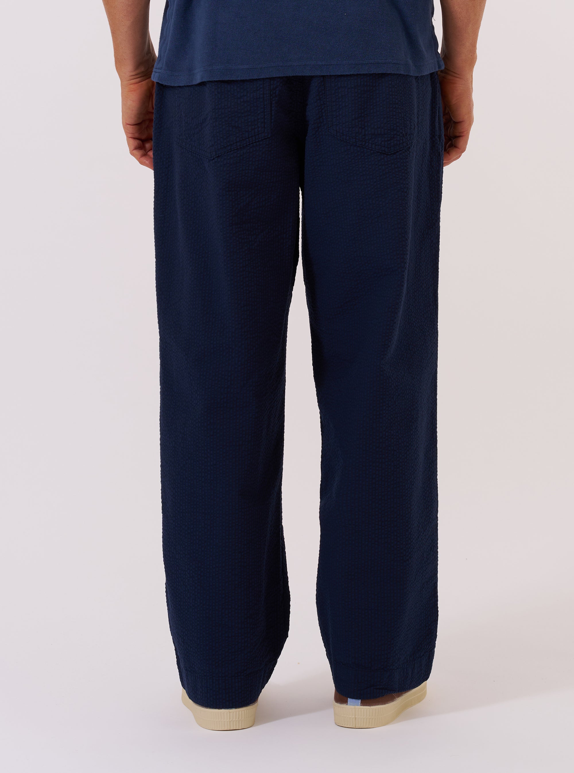 A model wears the Navy Derby Stripe Oxford Pant, shown from a rear three-quarter angle against a plain, neutral studio background. The trousers feature a loose, wide-leg fit and a navy and white vertical stripe pattern. The image highlights the clean back construction and the drape of the striped cotton fabric.