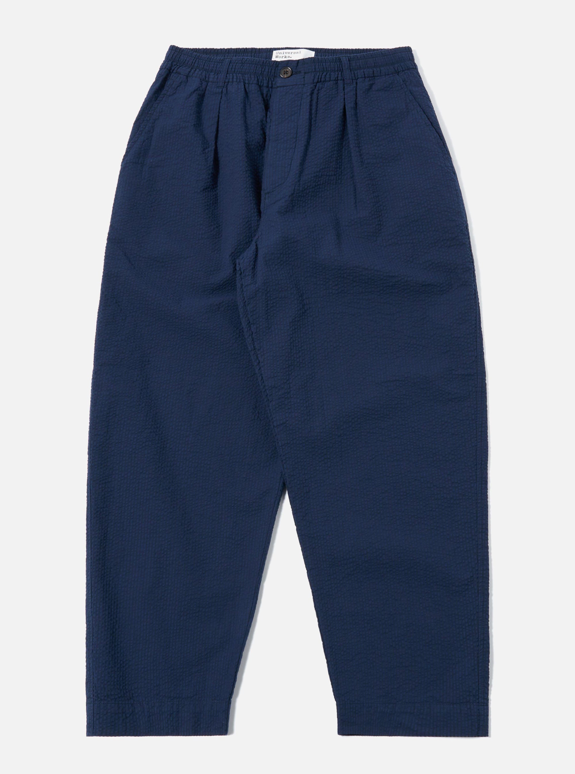 The Navy Derby Stripe Oxford Pant is shown as a flat lay against a plain white background. The trousers feature a vertical navy and white stripe pattern, a wide-leg fit, and double pleats at the waist. The design includes an elasticated waistband with an external drawstring and two side pockets.