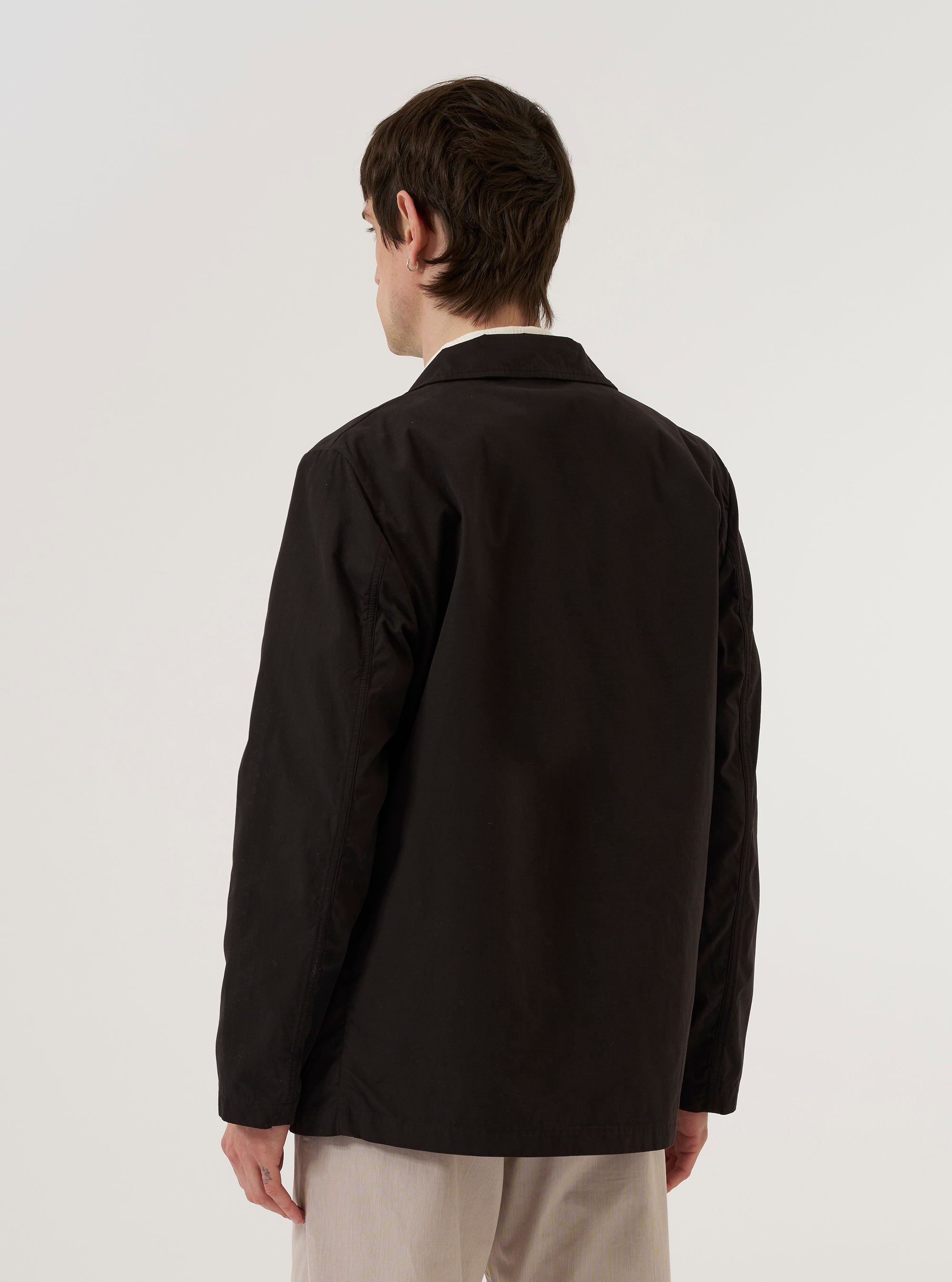 A model wears the Licorice Summer Canvas Two Button Jacket over a light-colored shirt, shown from a rear three-quarter angle against a plain white studio background. The jacket features a structured back, a single rear vent, and a relaxed tailored fit. The licorice brown fabric has a visible canvas texture.