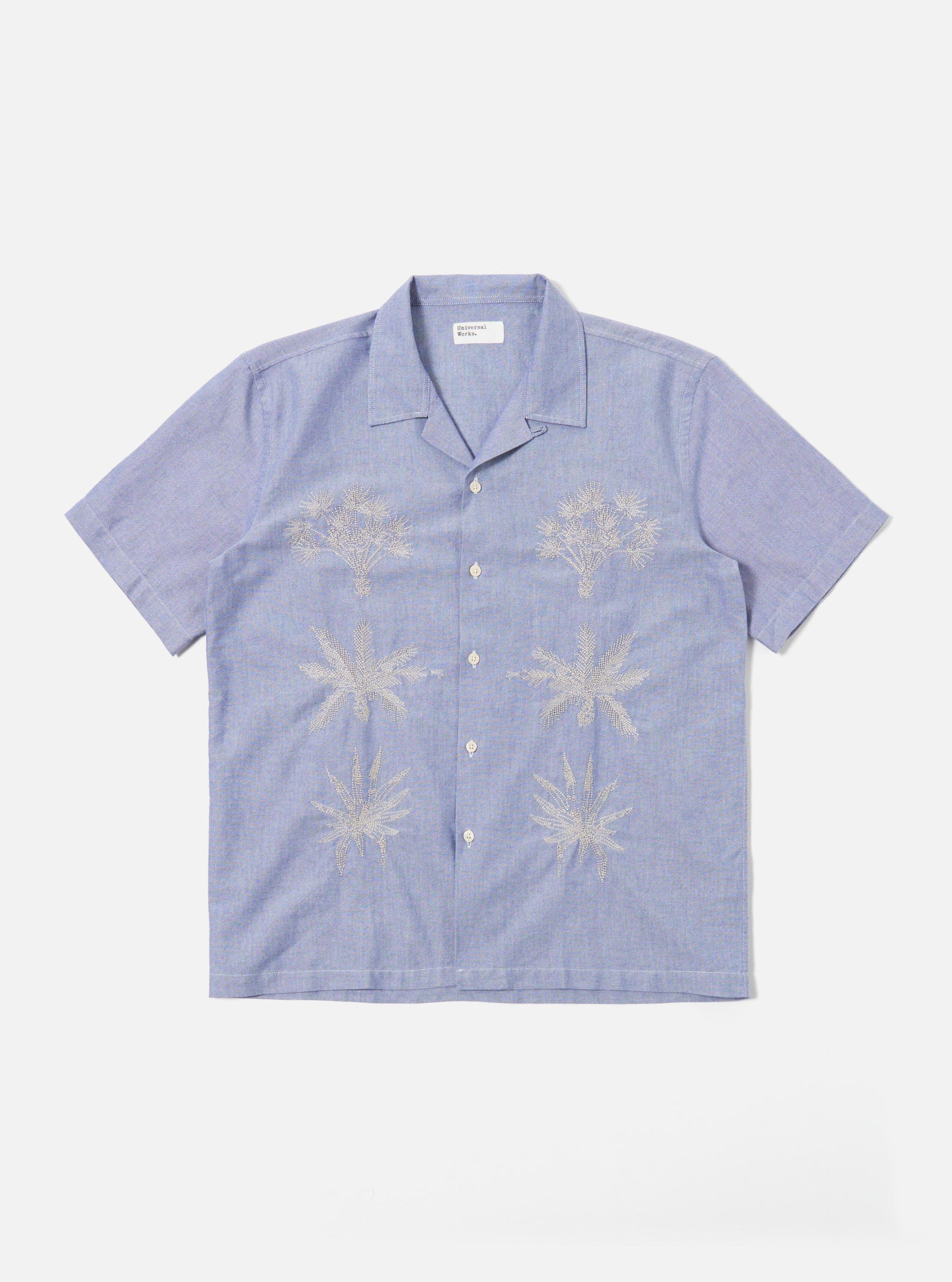 The Blue Oxford Shirting Road Trip Shirt is shown as a flat lay against a white background. The shirt features a camp collar, a central button-down front with tonal buttons, and a straight hem. The light blue cotton Oxford fabric has a visible weave, and a single patch pocket is located on the left chest.