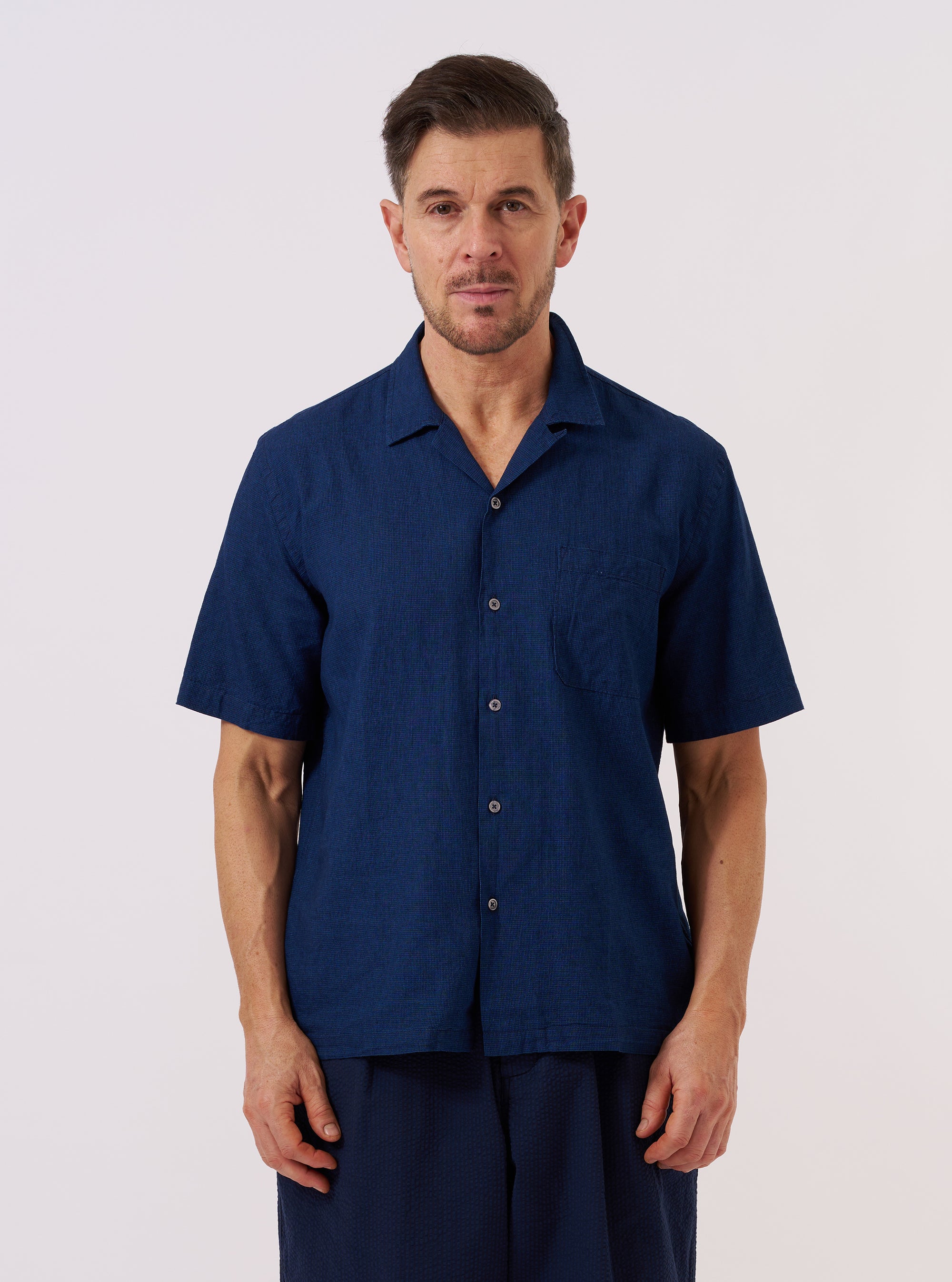 The Indigo Lincot Denim Road Shirt is worn by a model standing against a plain white background. The short-sleeve shirt is shown from the front, featuring a single chest patch pocket and a one-piece collar worn open. The model pairs the indigo-colored shirt with light-colored trousers.