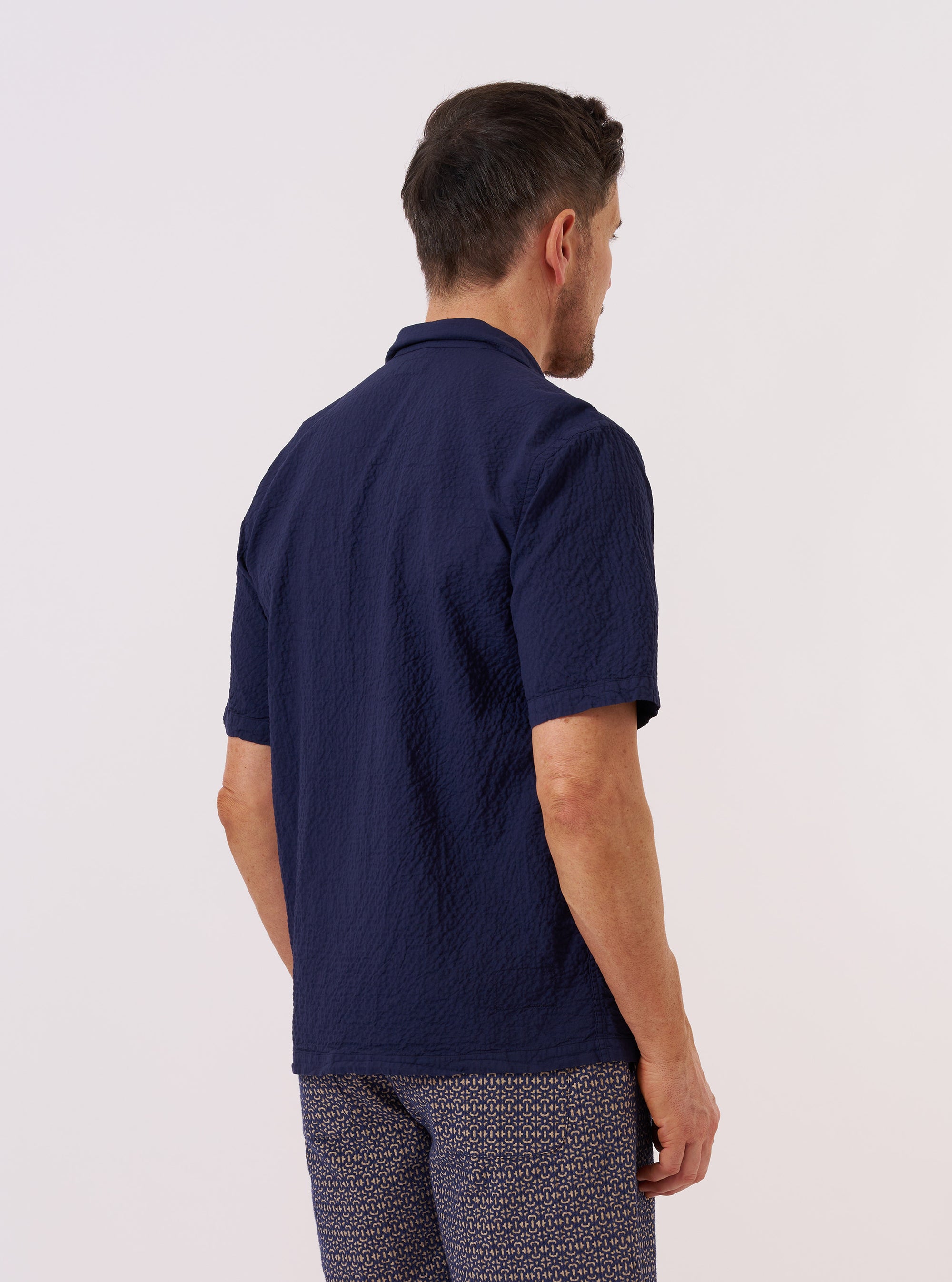 A model is wearing the Navy Arten Seersucker Road Shirt, shown from the back against a plain white background. The shirt features a relaxed fit, short sleeves, and a straight hem. The deep navy blue fabric has a distinct puckered seersucker texture, and the clean construction of the back panel is prominent. No mannequin is present in the image.