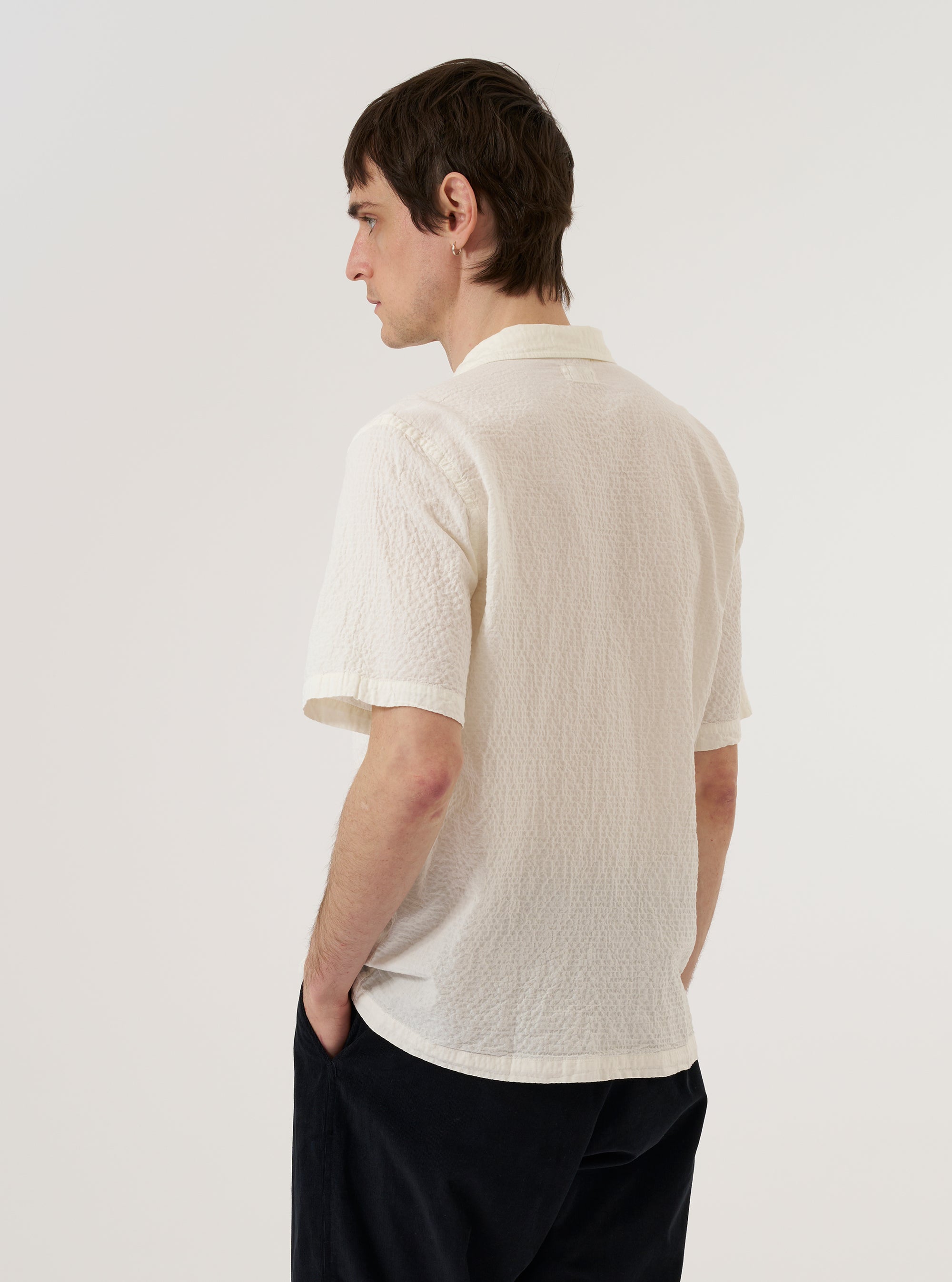 A model wears the Ecru Arten Seersucker Road Shirt against a plain white background. The shirt is shown from a rear three-quarter angle, highlighting its regular fit, straight hem, and short sleeves. The ecru-colored organic cotton fabric features a distinct puckered seersucker texture. No mannequin is present in the image.