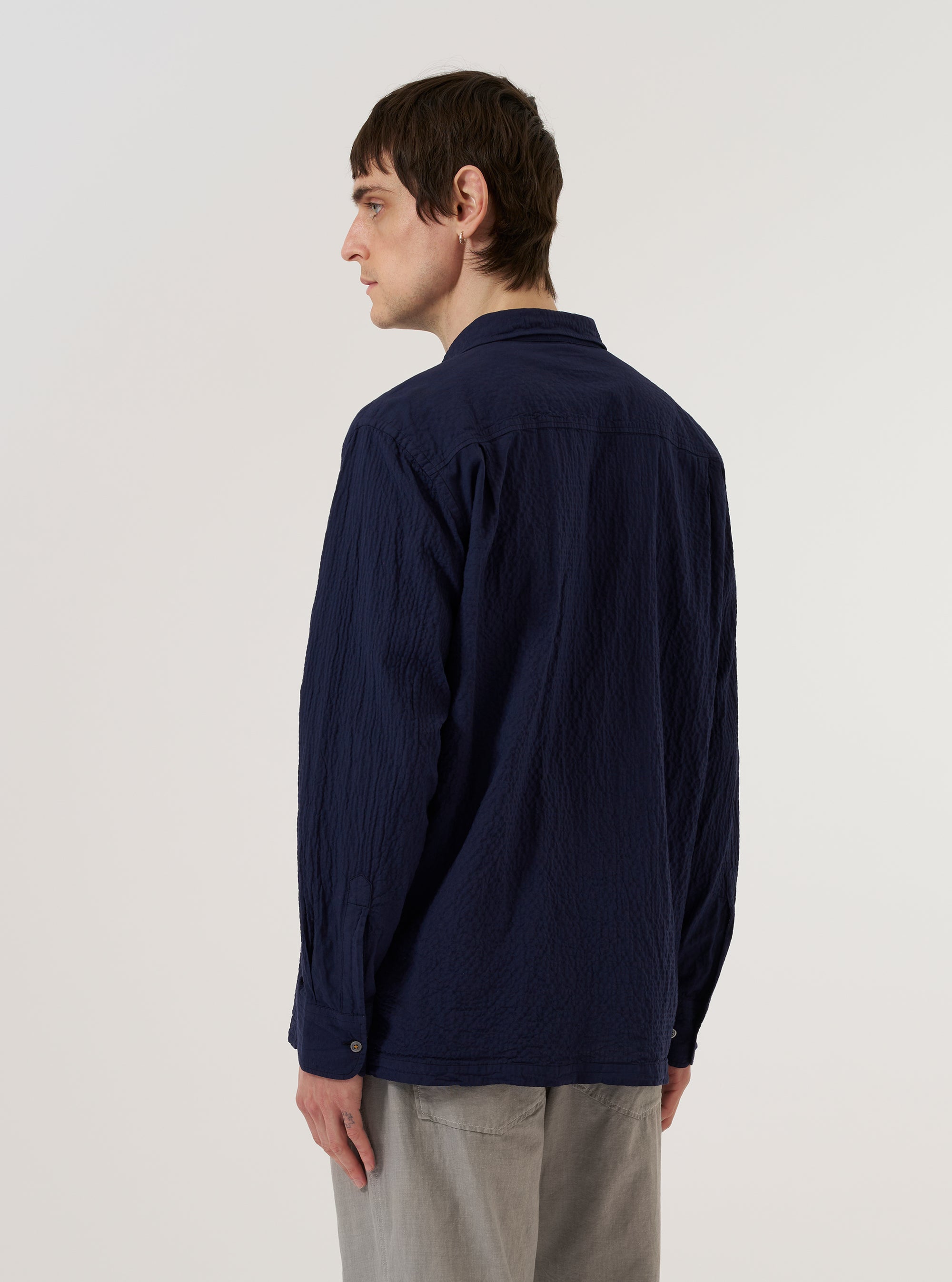 A model wears the Navy Arten Seersucker Attendant Shirt over a white t-shirt, shown from the rear three-quarter angle against a plain white background. The shirt features a loose fit, a straight hem, and a one-piece flat collar. The navy blue fabric has a distinct puckered seersucker texture.