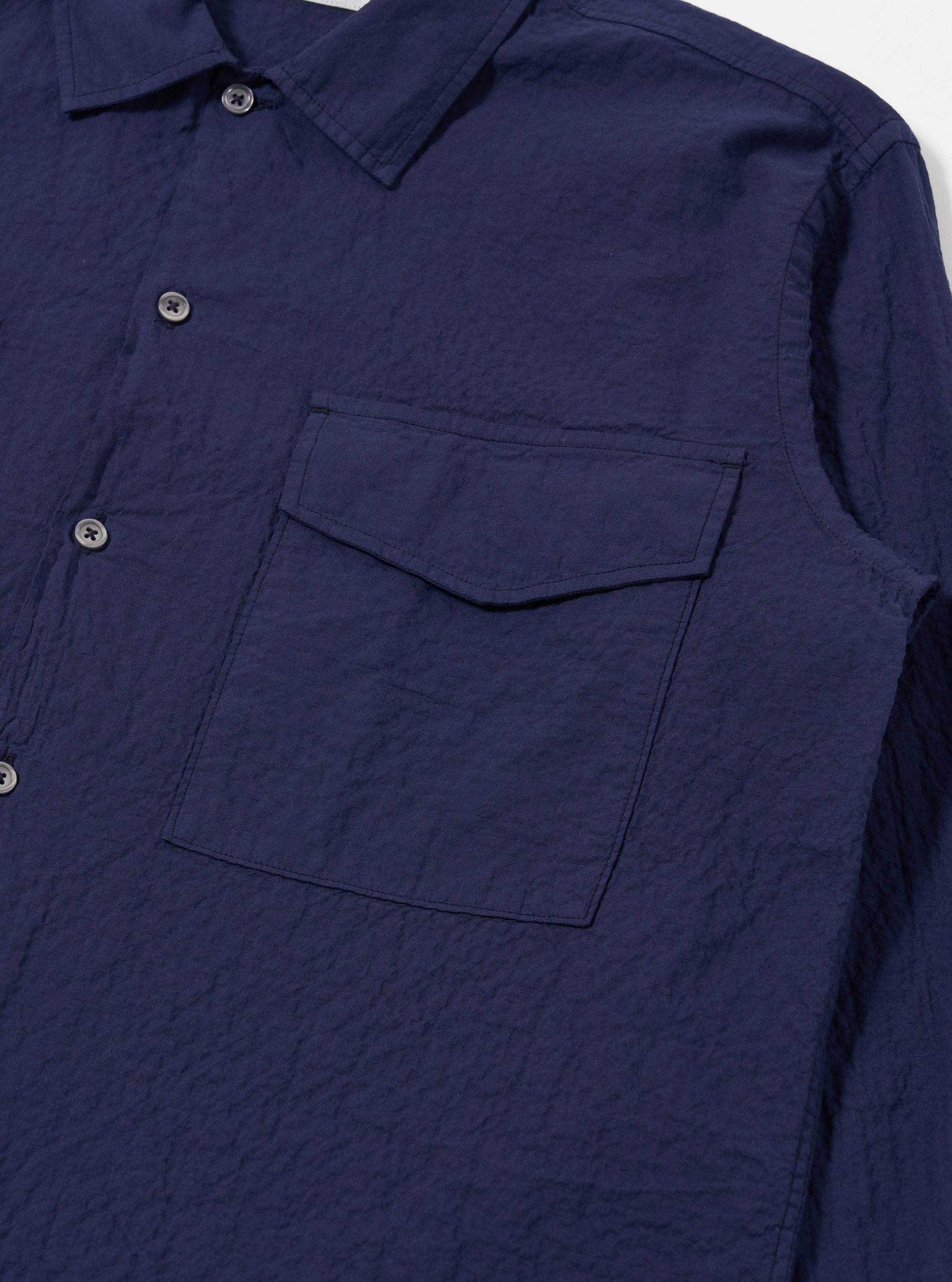 The Navy Arten Seersucker Attendant Shirt is shown from a rear angle as a flat lay against a plain white background. The image highlights the shirt's textured seersucker fabric, a straight hem, and a simple back yoke with a central box pleat.