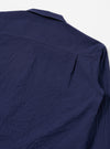 The Navy Arten Seersucker Attendant Shirt is worn by a model against a plain white background. The shirt features a five-button front closure, a simple pointed collar, and three patch pockets, including one at the chest and two at the waist. The dark navy organic cotton fabric has a distinct puckered seersucker texture. The model is shown from a front angle, wearing the shirt buttoned over a light-colored base layer. No mannequin is present in the image.