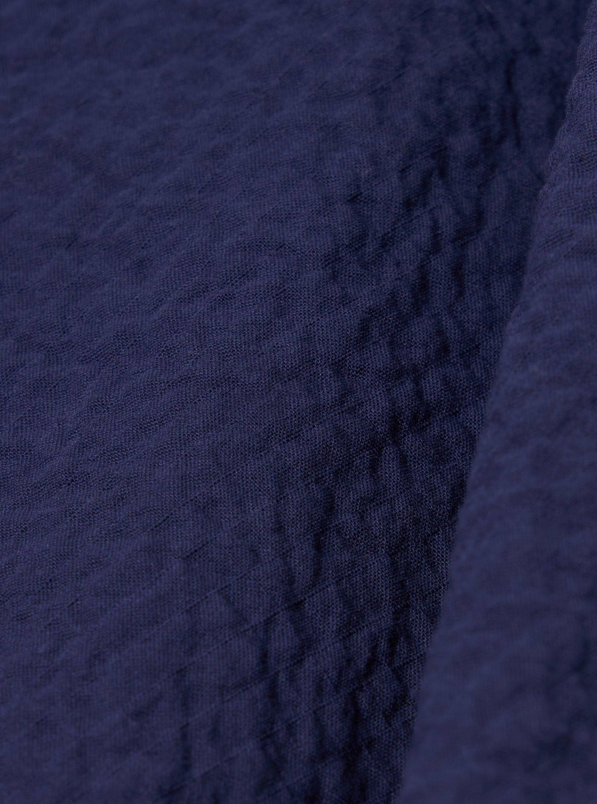 A close-up fabric swatch of the Navy Arten Seersucker Attendant Shirt highlights the deep navy blue color and the distinct puckered, crinkled texture of the organic cotton seersucker fabric. The material fills the entire frame, providing a detailed view of the textile's weave.
