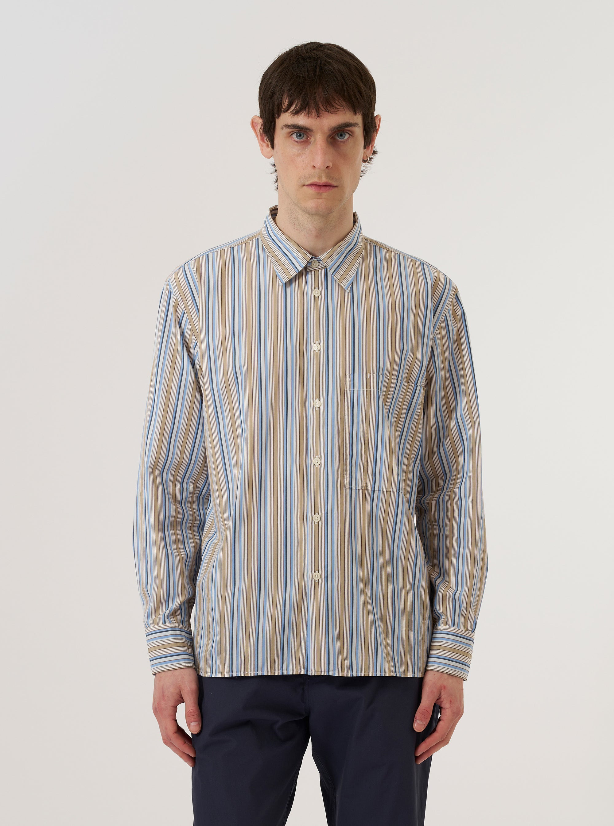A model wears the Multi Pop Multi Stripe Square Pocket Shirt against a plain white background. The shirt features a vibrant multi-colored vertical stripe pattern throughout, a central button-down front, a structured collar, and a prominent square patch pocket on the chest. The image captures the model from the front, highlighting the shirt's relaxed fit and the crisp texture of the poplin fabric.